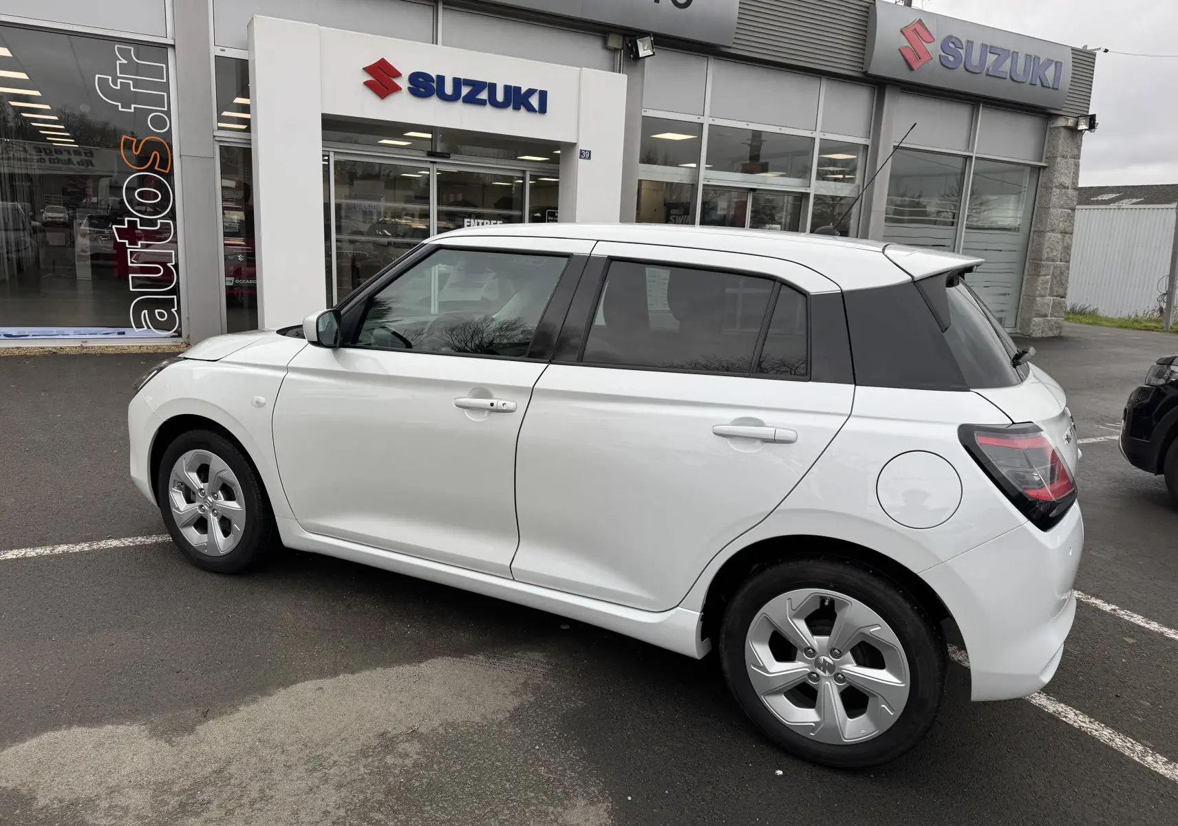 Vue côté droit arrière d'une Suzuki Swift 1.2 Privilège Hybrid blanche, avec vitres teintées et jantes alliage, devant un concessionnaire.