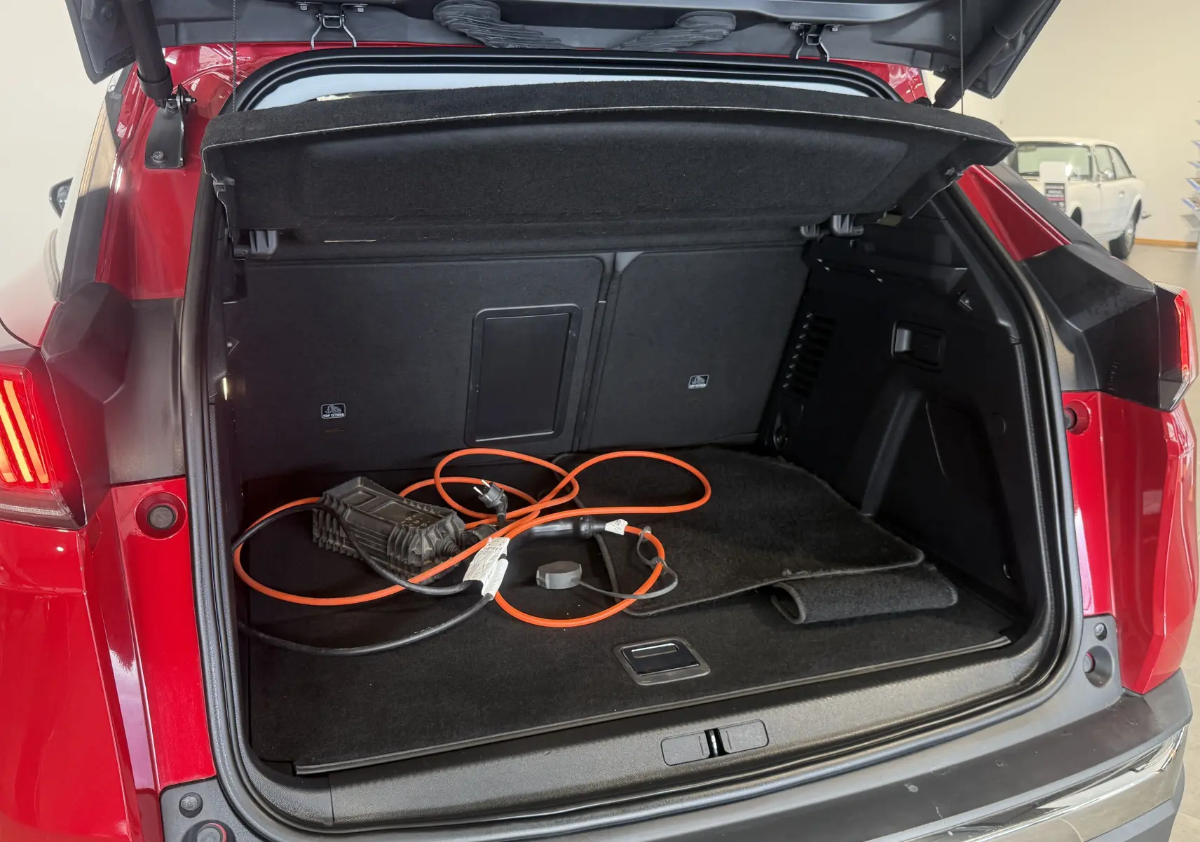 Coffre ouvert vu de l’arrière d’un Peugeot 3008 Hybrid rouge 2025 avec câble de recharge orange visible.