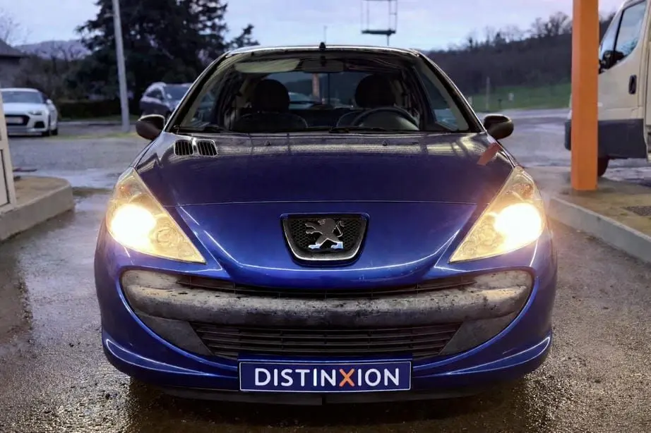 Peugeot 206+ bleu vue 3/4 avant droit avec phares allumés, garée devant un bâtiment vitré.