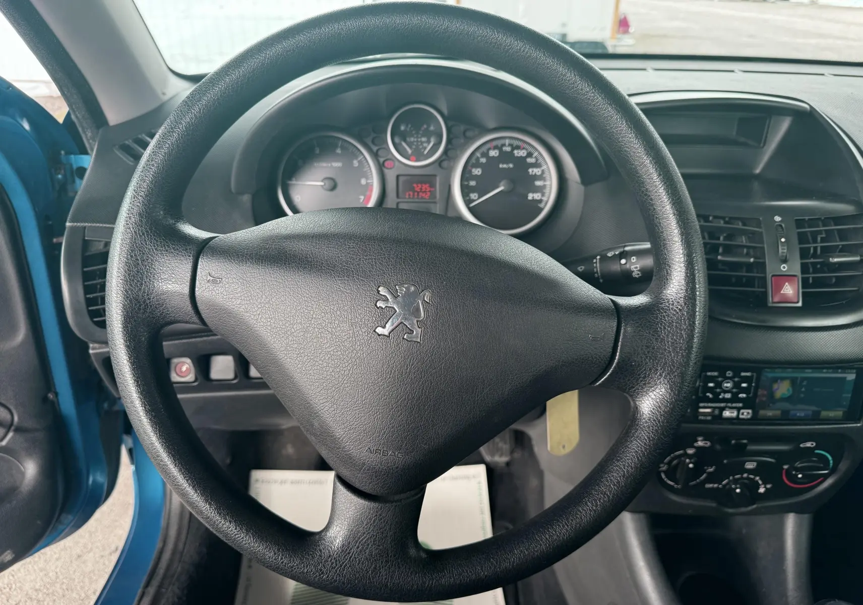 Gros plan sur les commandes noires côté gauche du tableau de bord d'une Peugeot 206+ bleue, avec réglage des phares et airbag passager.