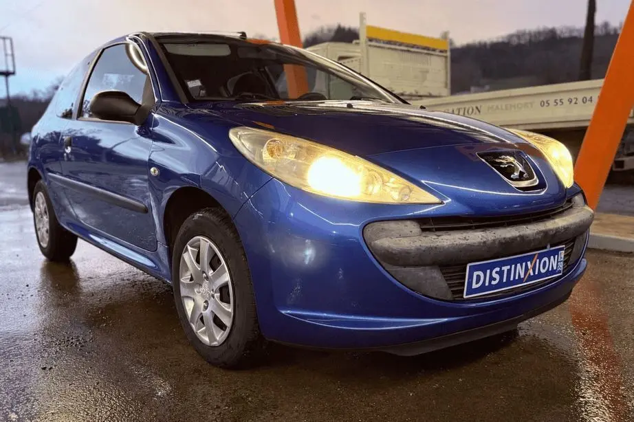 Peugeot 206+ bleu vue de profil côté gauche sous abri, phare avant allumé sur sol mouillé.