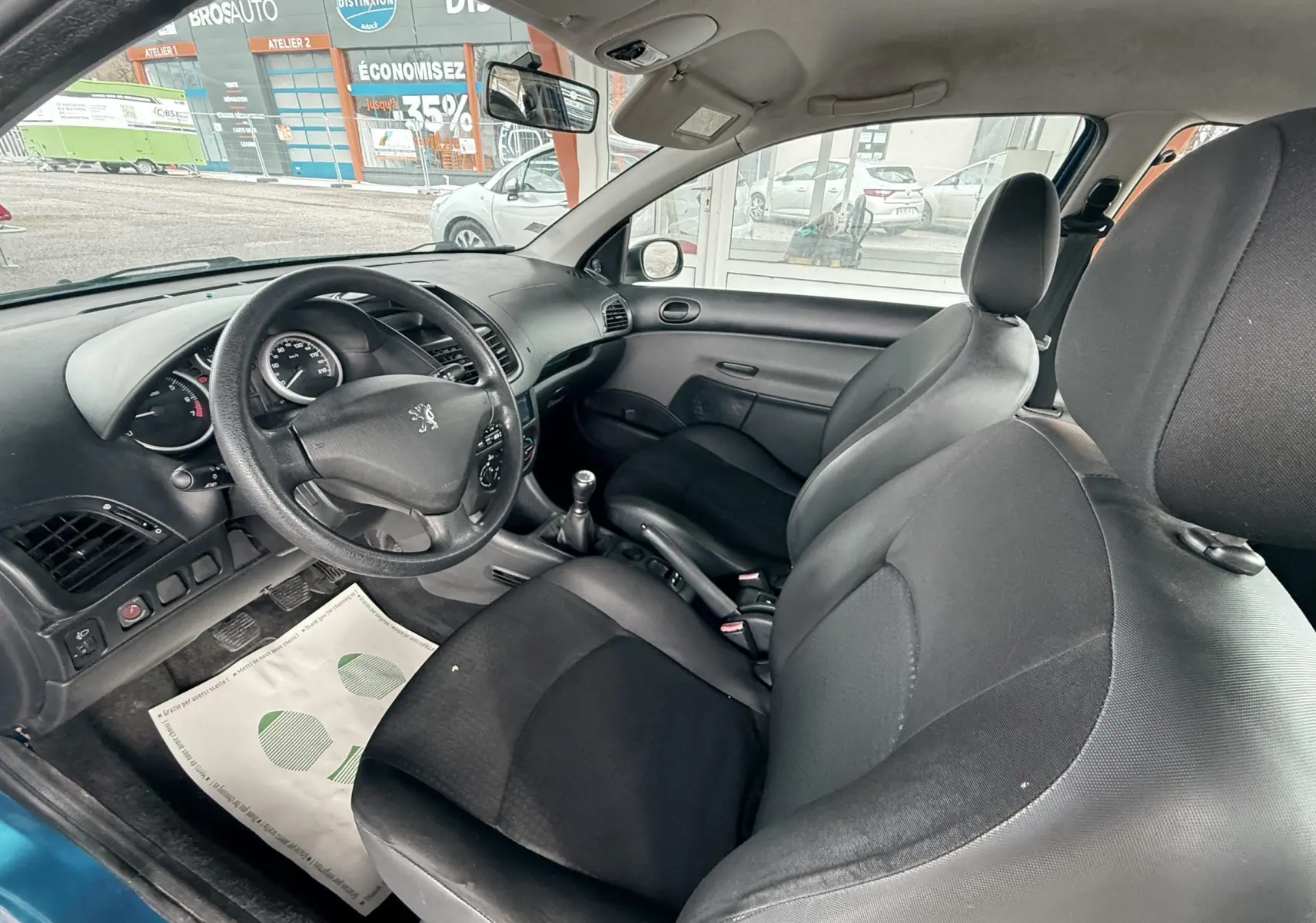 Peugeot 206+ bleu vue 3/4 avant droit avec phares allumés sur sol mouillé, devant structure orange.