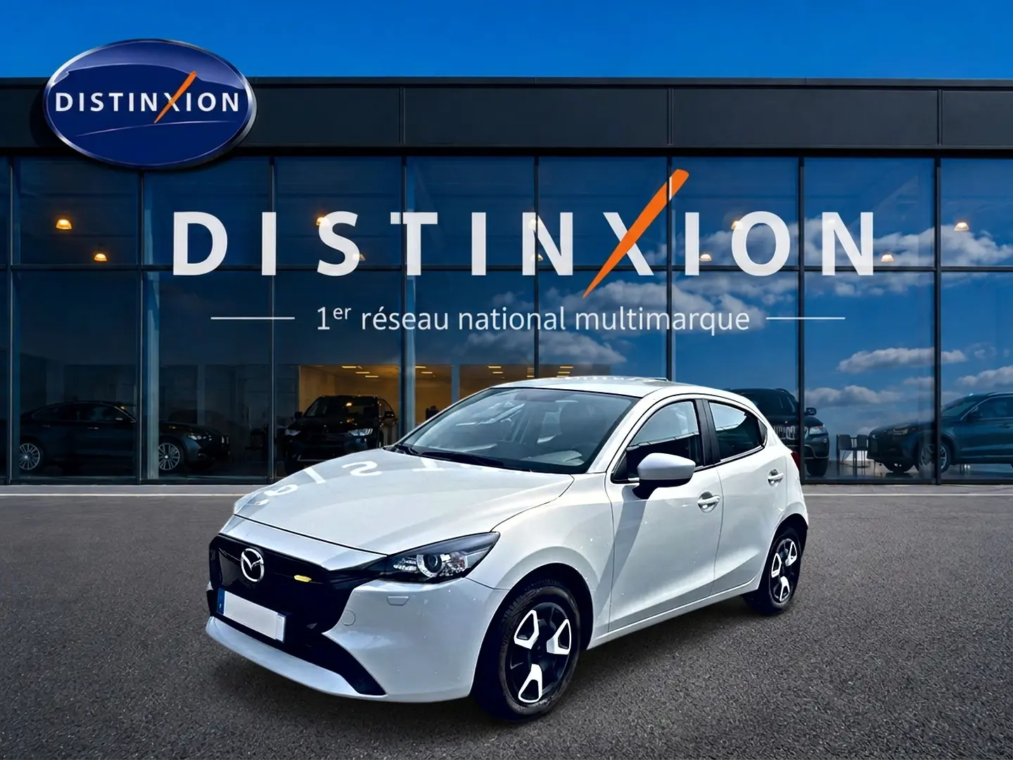 Vue 3/4 avant droite d'une Mazda2 blanche 2024 avec calandre noire et jantes au design distinctif, devant un showroom Distinxion.