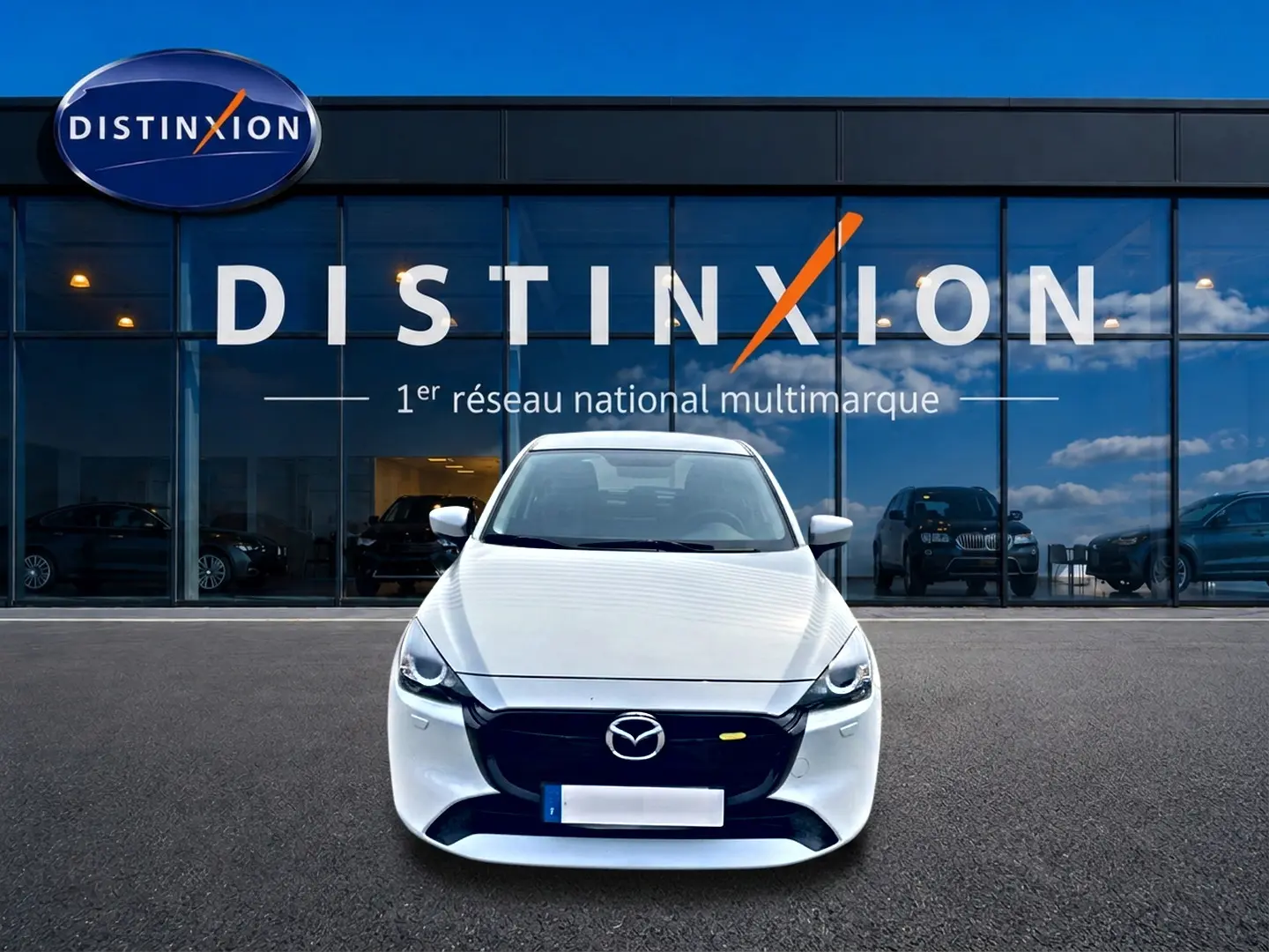 Vue frontale d'une Mazda2 blanche 2024 garée devant une vitrine avec logo Distinxion en arrière-plan.