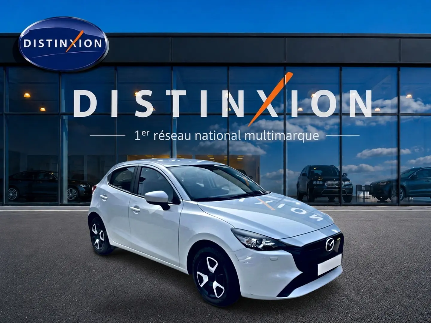Vue 3/4 avant gauche d'une Mazda2 blanche 2024 avec calandre noire et jantes distinctives devant un showroom Distinxion.