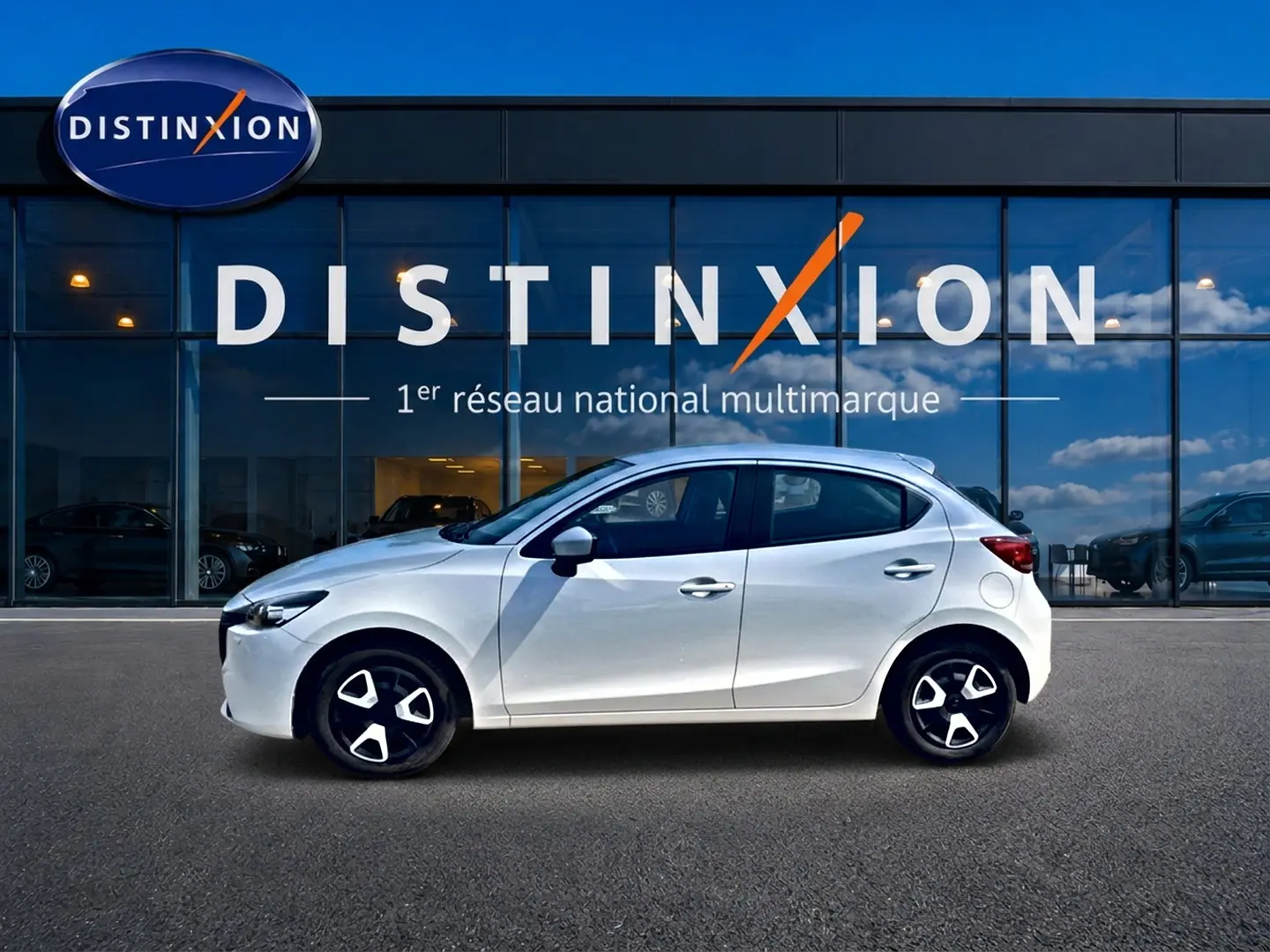Vue de profil côté gauche d'une Mazda2 blanche 2024 garée devant une vitrine avec logo Distinxion.