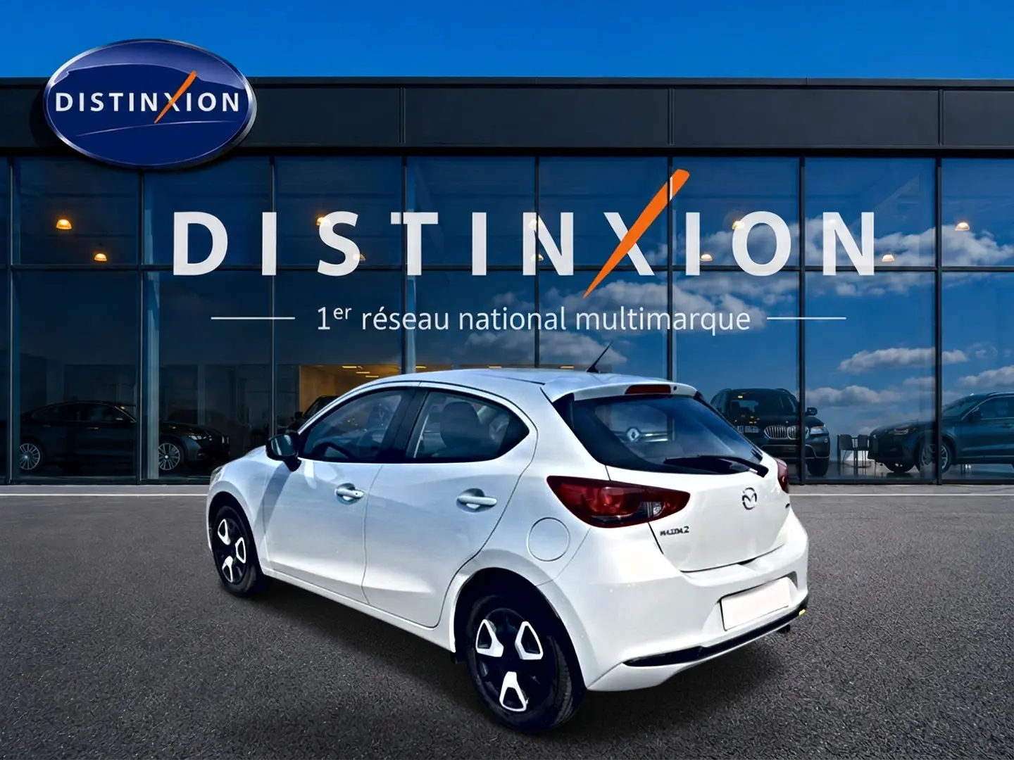 Vue 3/4 arrière droite d'une Mazda2 blanche 2024 avec jantes distinctives et vitres teintées, devant une vitrine Distinxion.