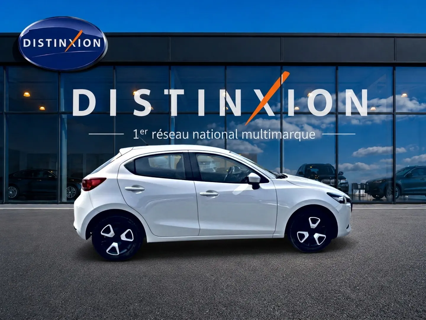 Vue latérale droite d'une Mazda2 blanche 2024 avec jantes noires et détails modernes, stationnée devant une vitrine Distinxion.