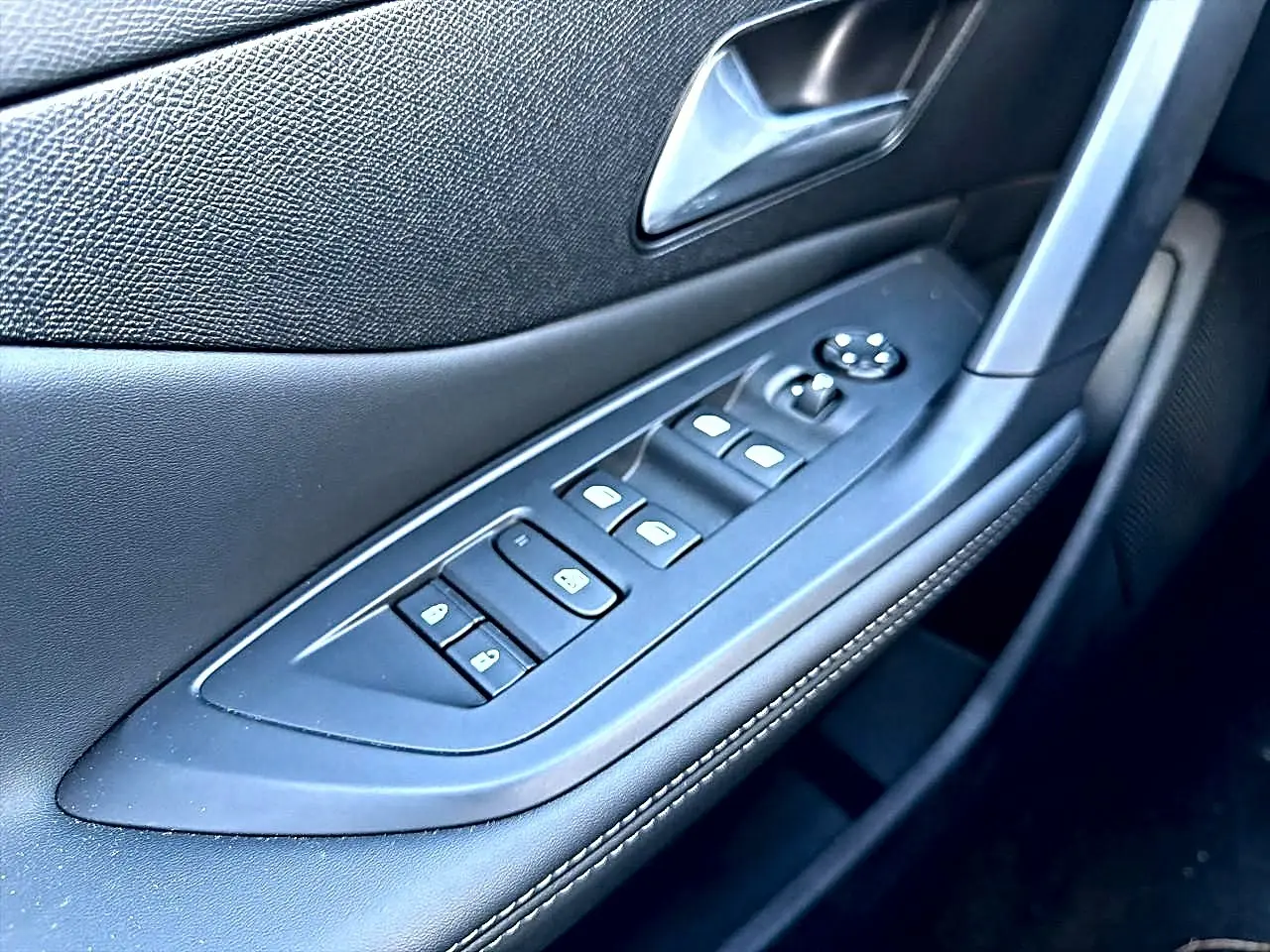 Gros plan sur la commande des vitres électriques et rétroviseurs côté conducteur en cuir noir avec surpiqûres blanches.