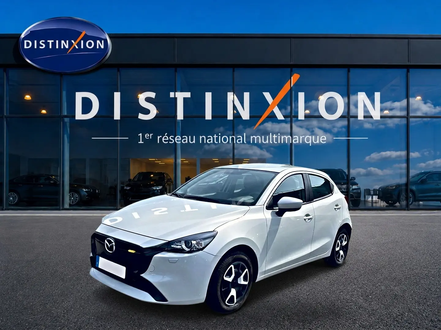 Vue 3/4 avant droite d'une Mazda2 blanche 2024 avec calandre noire et jantes au design moderne devant un showroom Distinxion.