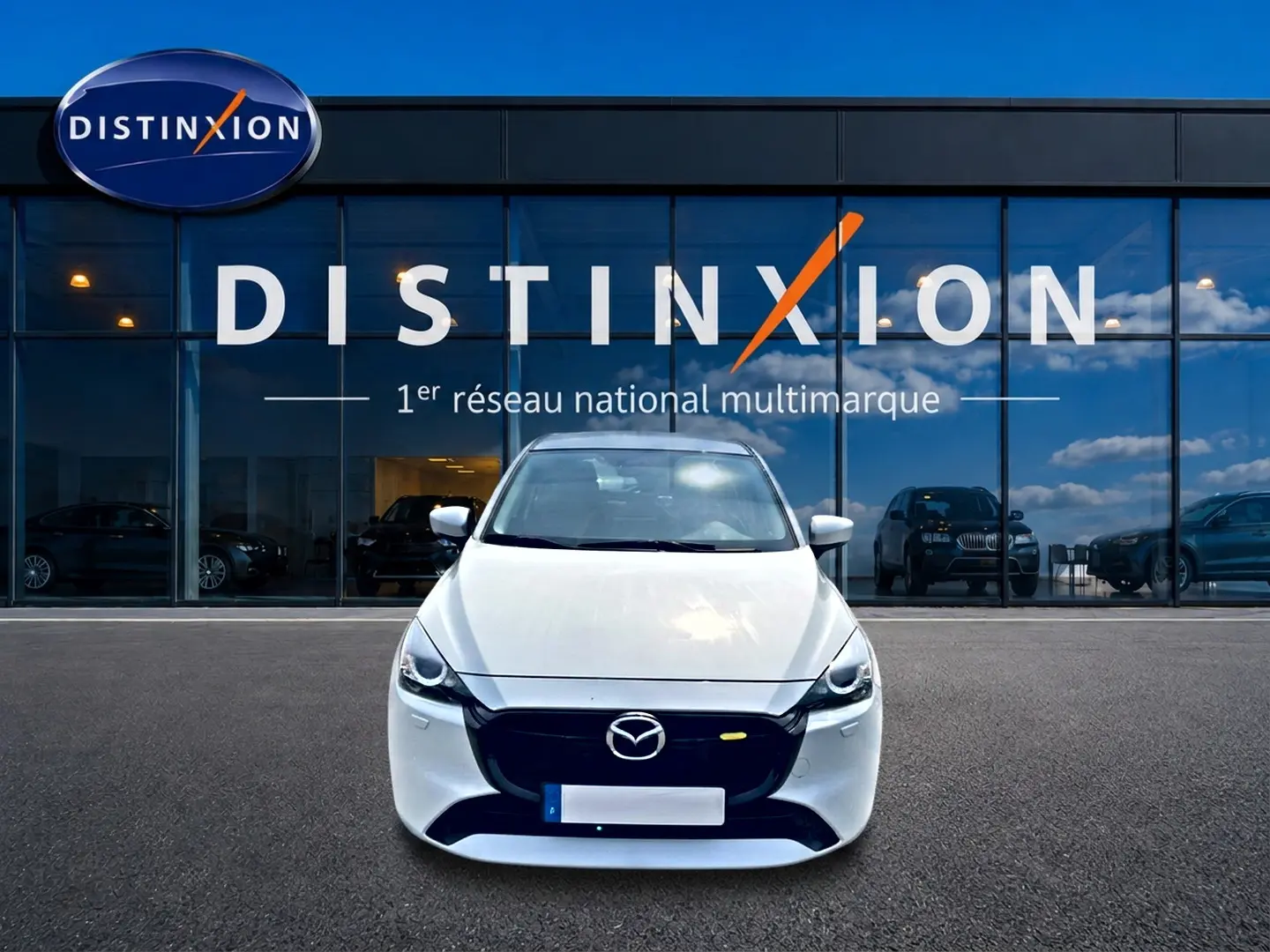 Vue avant d'une Mazda2 blanche 2024 avec calandre noire, stationnée devant une vitrine Distinxion.