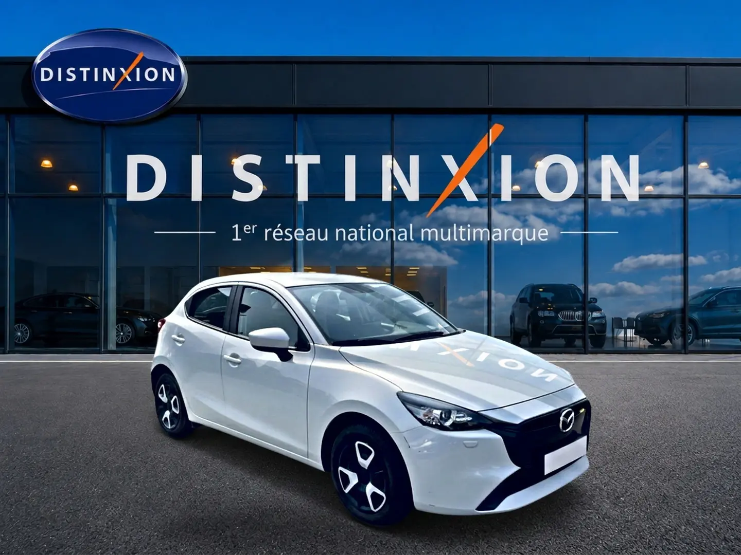 Vue 3/4 avant droite d'une Mazda2 blanche garée devant une vitrine avec logo Distinxion en arrière-plan.