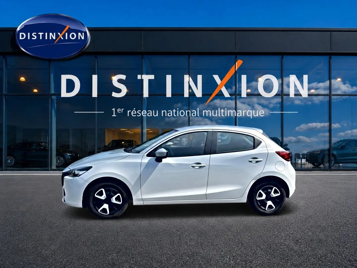 Vue latérale droite d'une Mazda2 blanche 2024 garée devant une vitrine avec logo Distinxion.