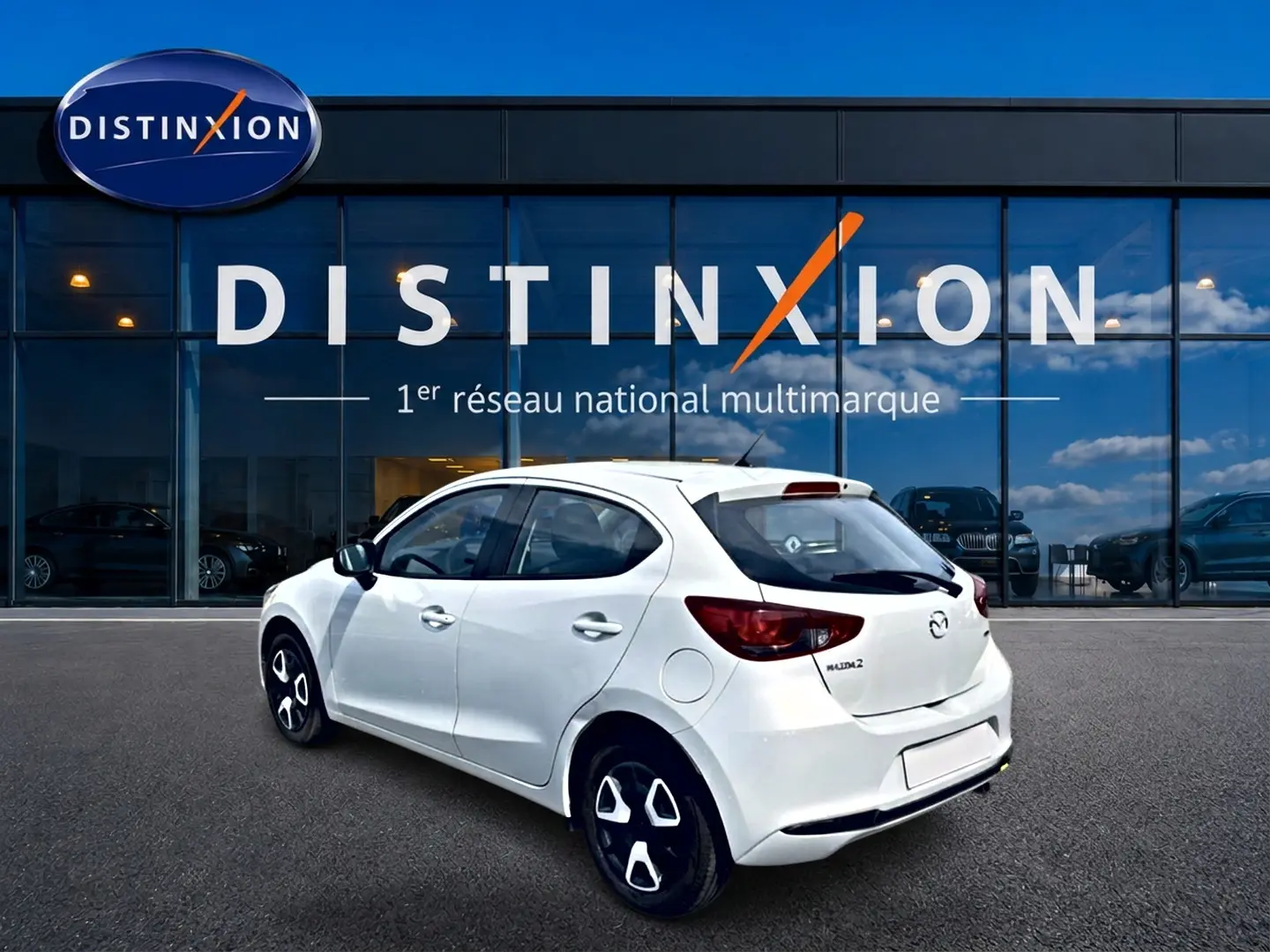Vue 3/4 arrière droite d'une Mazda2 blanche 2024 avec jantes distinctives, garée devant une vitrine Distinxion.