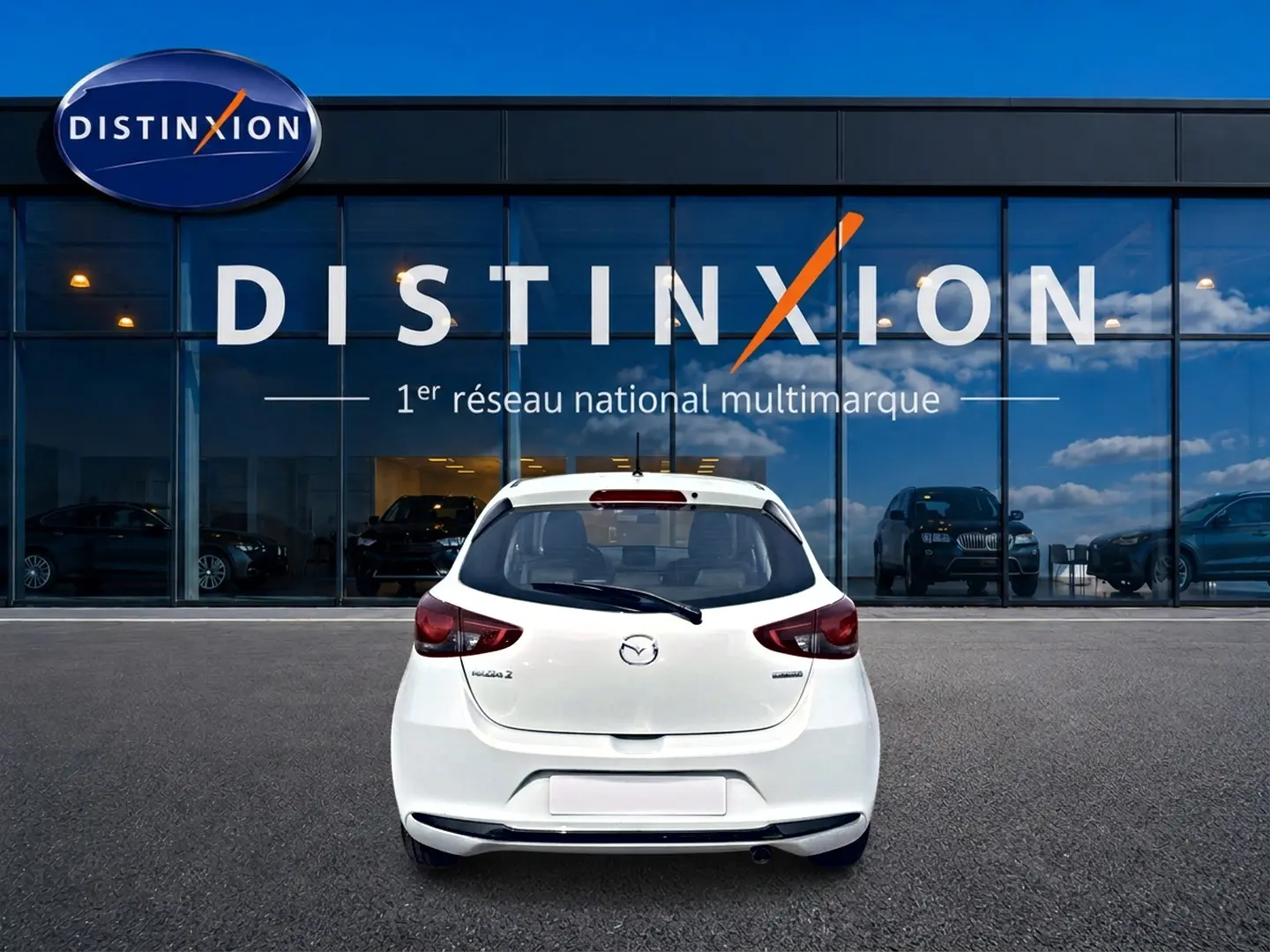Vue arrière d'une Mazda2 blanche 2024 garée devant une vitrine avec logo Distinxion en arrière-plan.