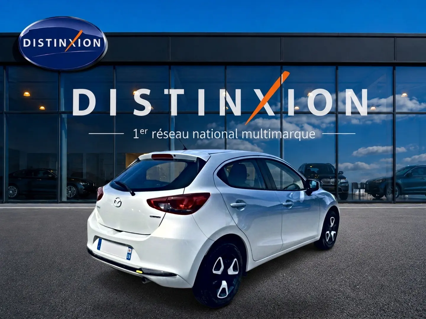 Vue 3/4 arrière droite d'une Mazda2 blanche 2024 garée devant une vitrine avec logo Distinxion.