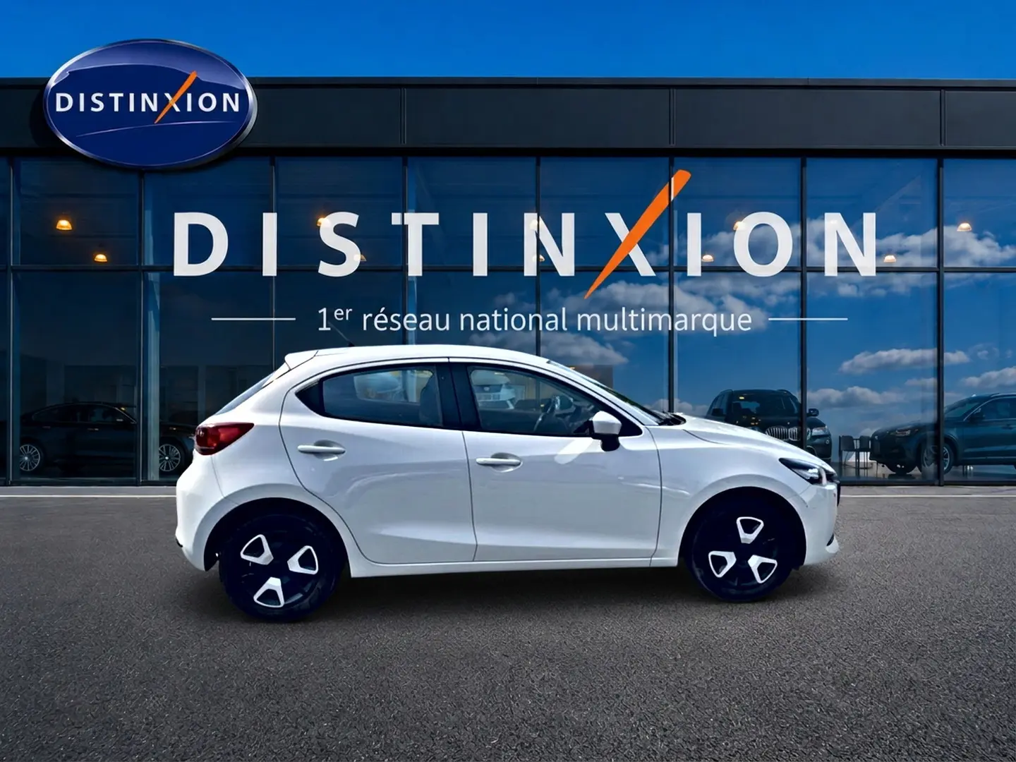 Vue de profil côté gauche d'une Mazda2 blanche 2024 garée devant une vitrine avec logo Distinxion.