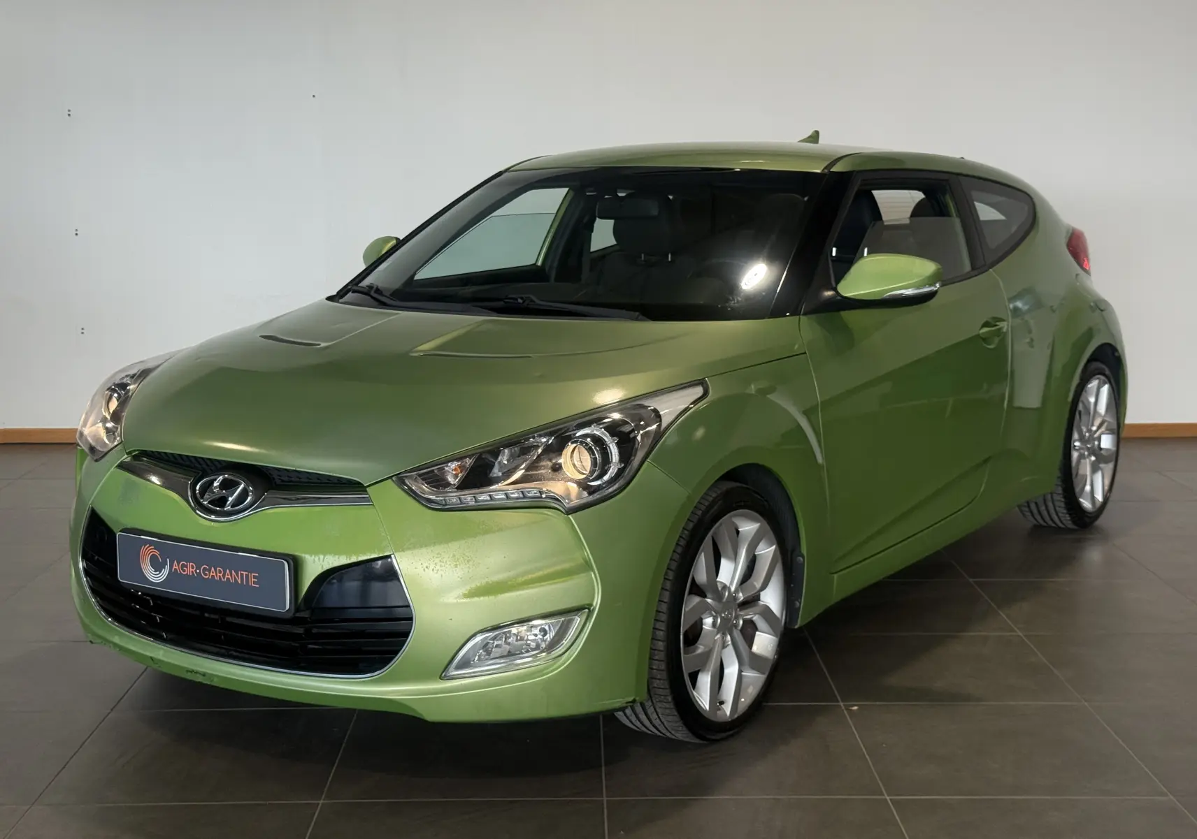 Hyundai Veloster vert 2025 en 3/4 avant droit, avec phares allumés et jantes alliage visibles.
