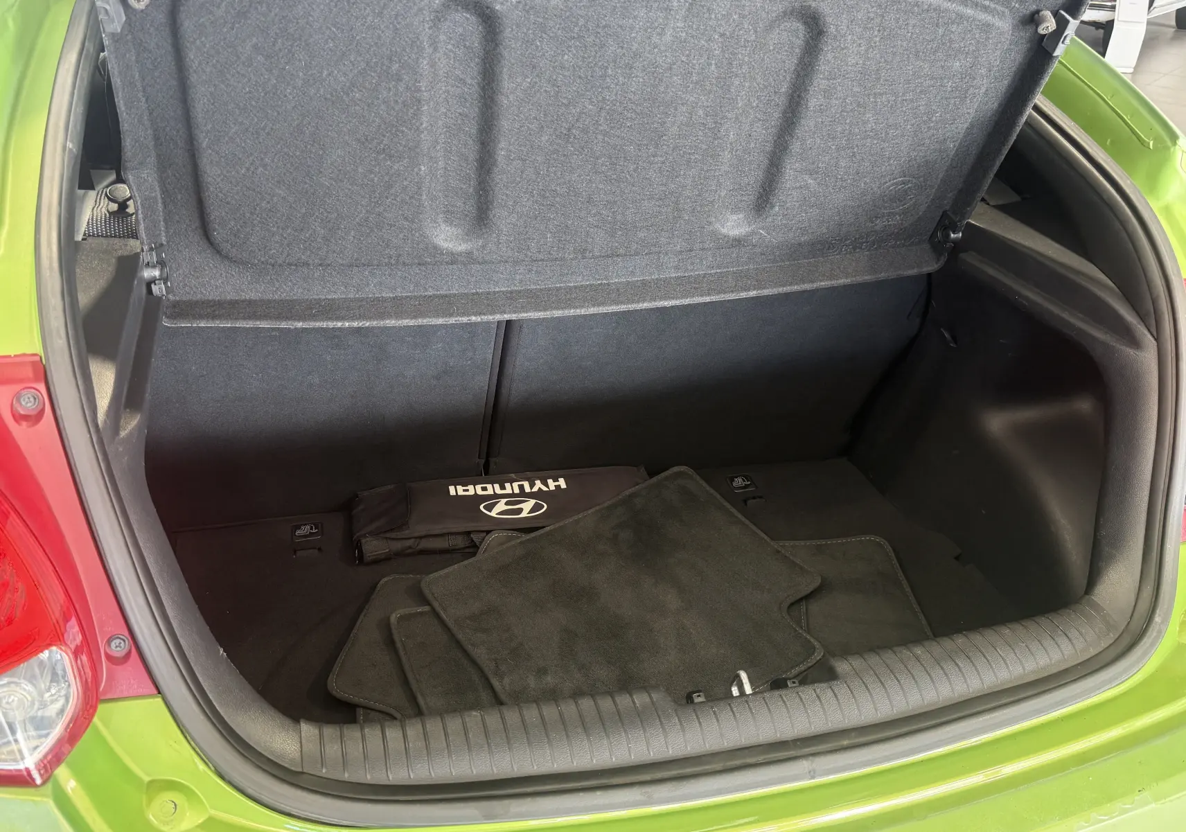 Coffre ouvert vu de l'arrière d'une Hyundai Veloster 2025 vert, avec tapis de sol et trousse Hyundai visibles à l'intérieur.