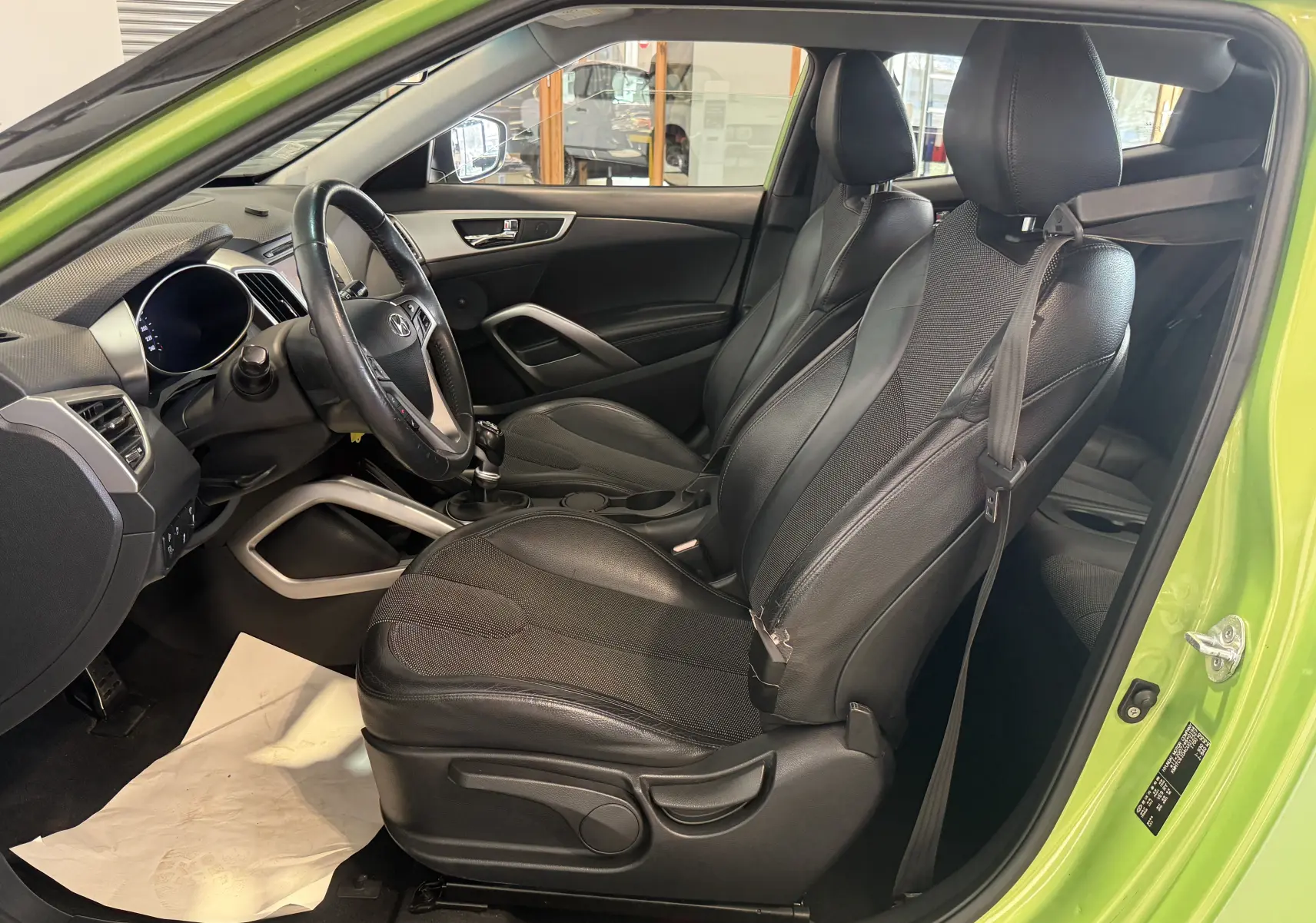 Vue intérieure côté gauche de la Hyundai Veloster 2025 verte, montrant les sièges noirs et le volant avec boîte manuelle.