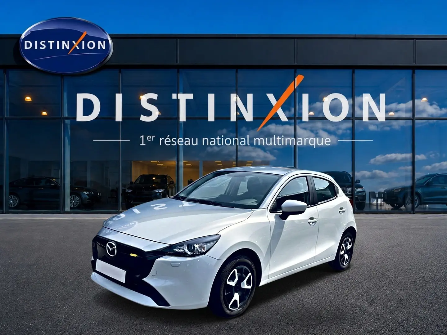 Vue 3/4 avant droite d'une Mazda2 blanche 2024 avec jantes noires et calandre noire devant un showroom Distinxion.