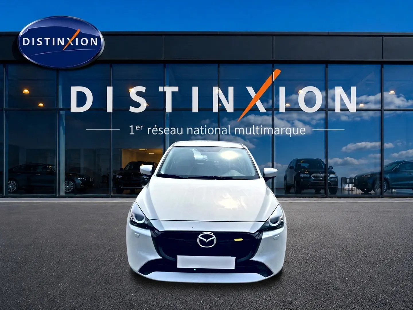 Vue avant d'une Mazda2 blanche 2024 stationnée devant une vitrine avec logo Distinxion en arrière-plan.