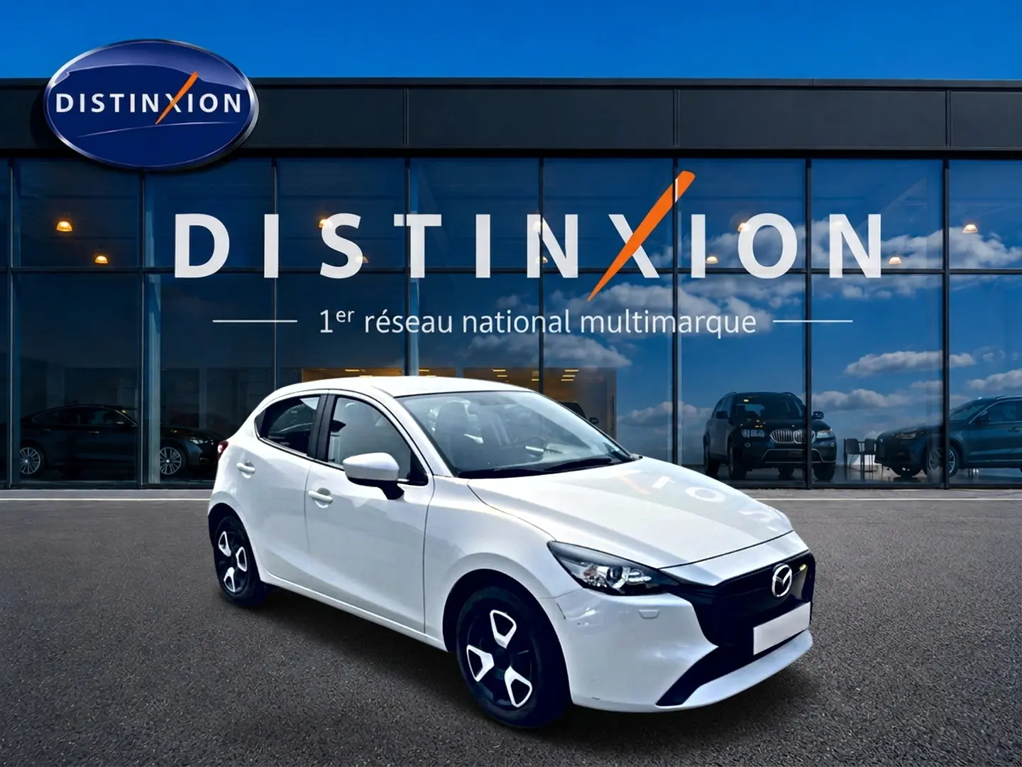 Vue 3/4 avant droite d'une Mazda2 blanche 2024 avec jantes noires et calandre noire devant un showroom Distinxion.