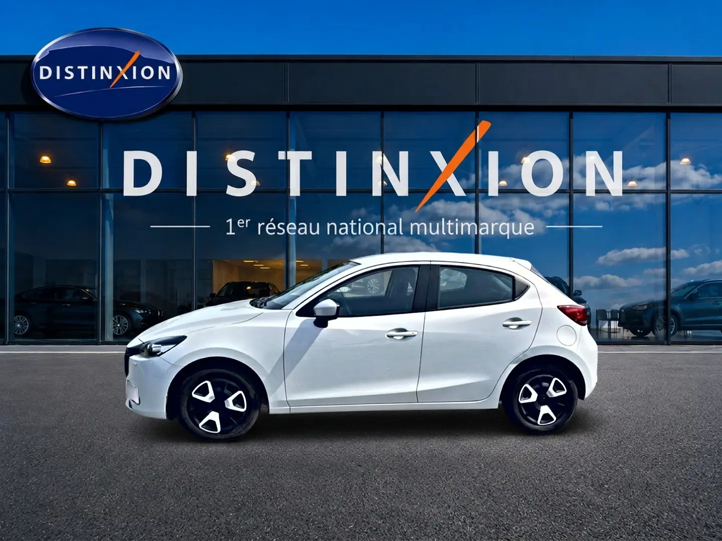 Vue de profil côté gauche d'une Mazda2 blanche 2024 garée devant une vitrine avec logo Distinxion.