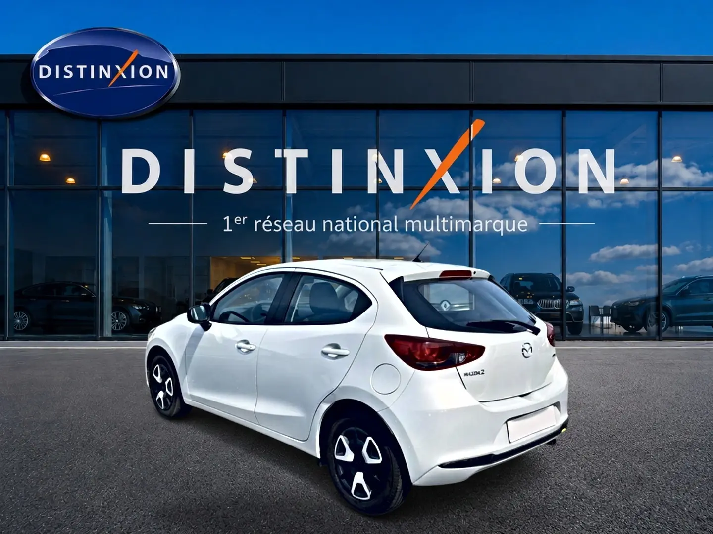 Vue 3/4 arrière droite d'une Mazda2 blanche 2024 avec jantes noires et feux arrière foncés, devant une vitrine Distinxion.