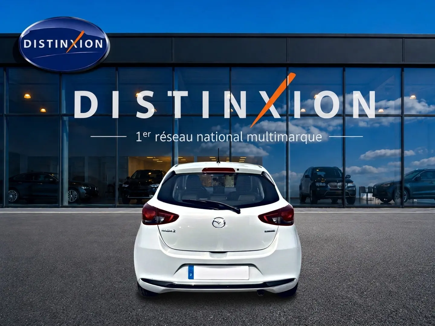 Vue arrière d'une Mazda2 blanche 2024 garée devant une vitrine avec logo Distinxion en arrière-plan.