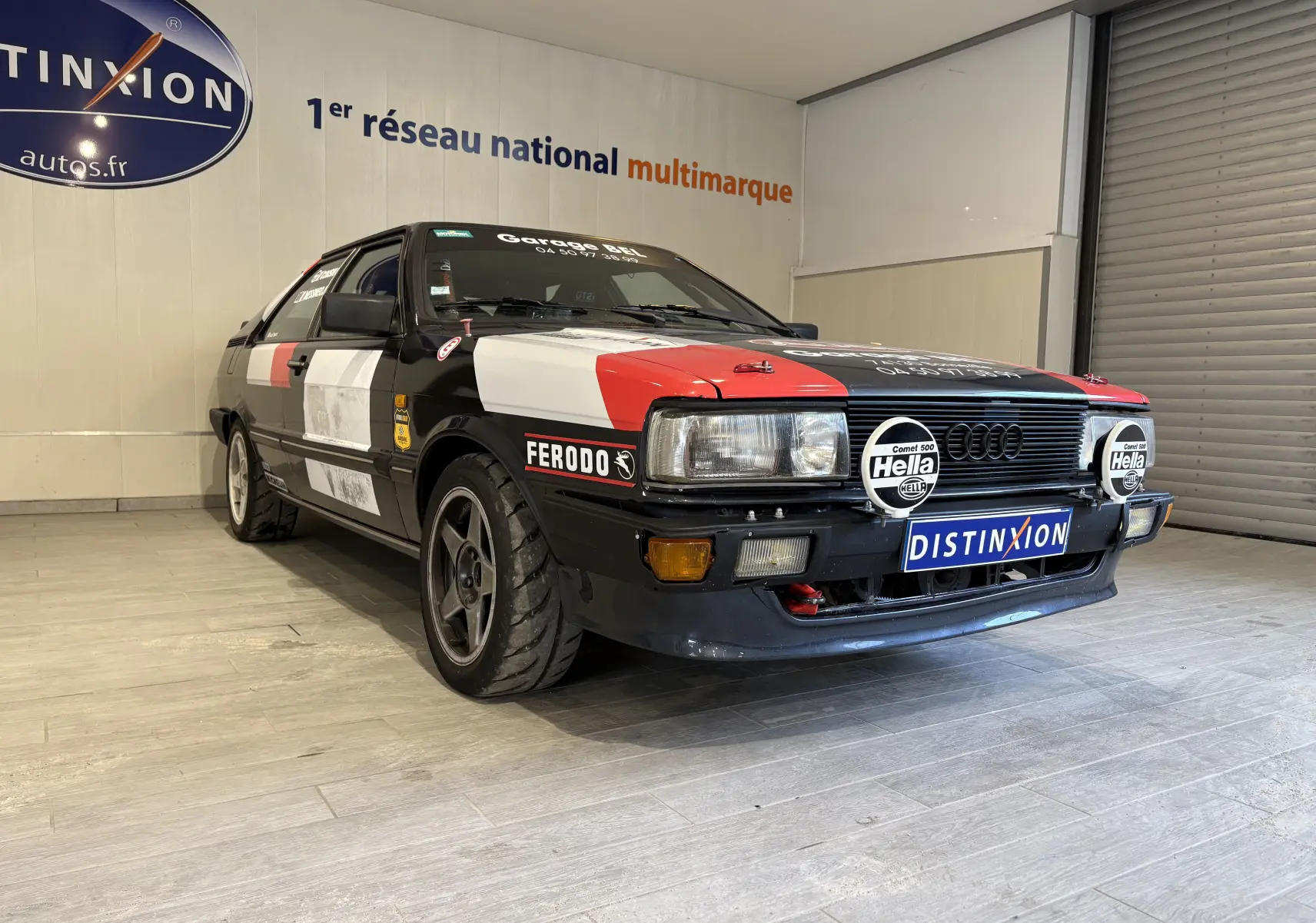 Vue frontale d'une Audi Coupé GT noire avec capot rouge, équipée de deux phares additionnels Hella et stickers de rallye.