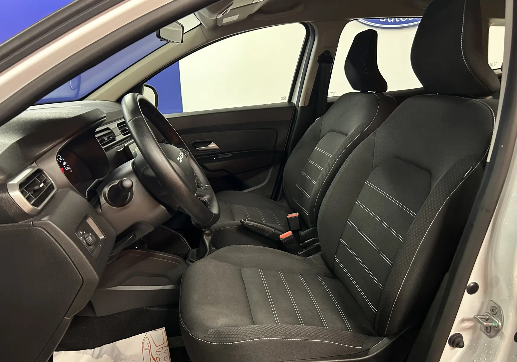 Vue intérieure côté conducteur du Dacia Duster blanc 2025, montrant les sièges en tissu noir et le volant multifonction.