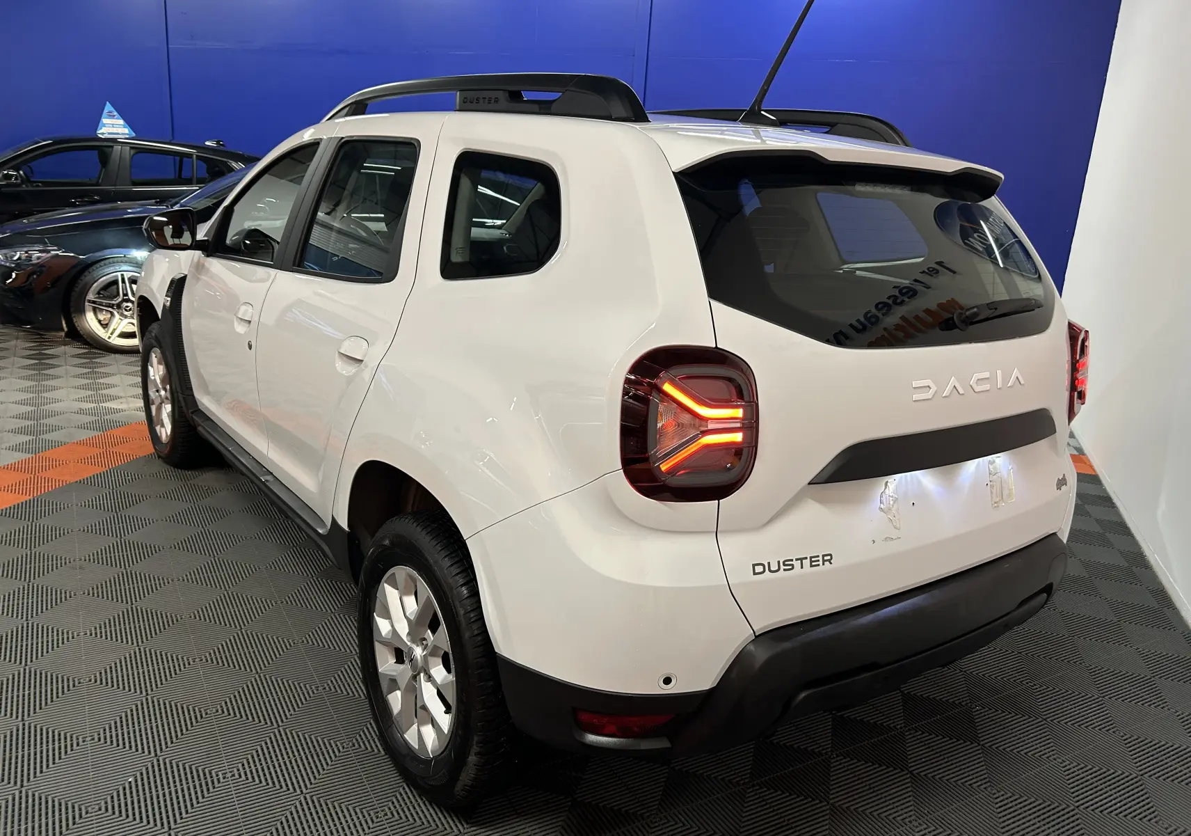 Vue 3/4 arrière droite du Dacia Duster blanc 2025 avec feux arrière allumés et toit noir en showroom.