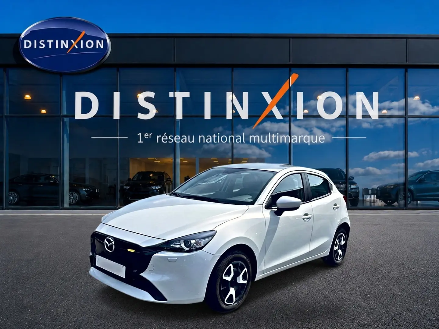Vue 3/4 avant droite d'une Mazda2 blanche 2024 avec calandre noire et jantes distinctives devant un showroom Distinxion.
