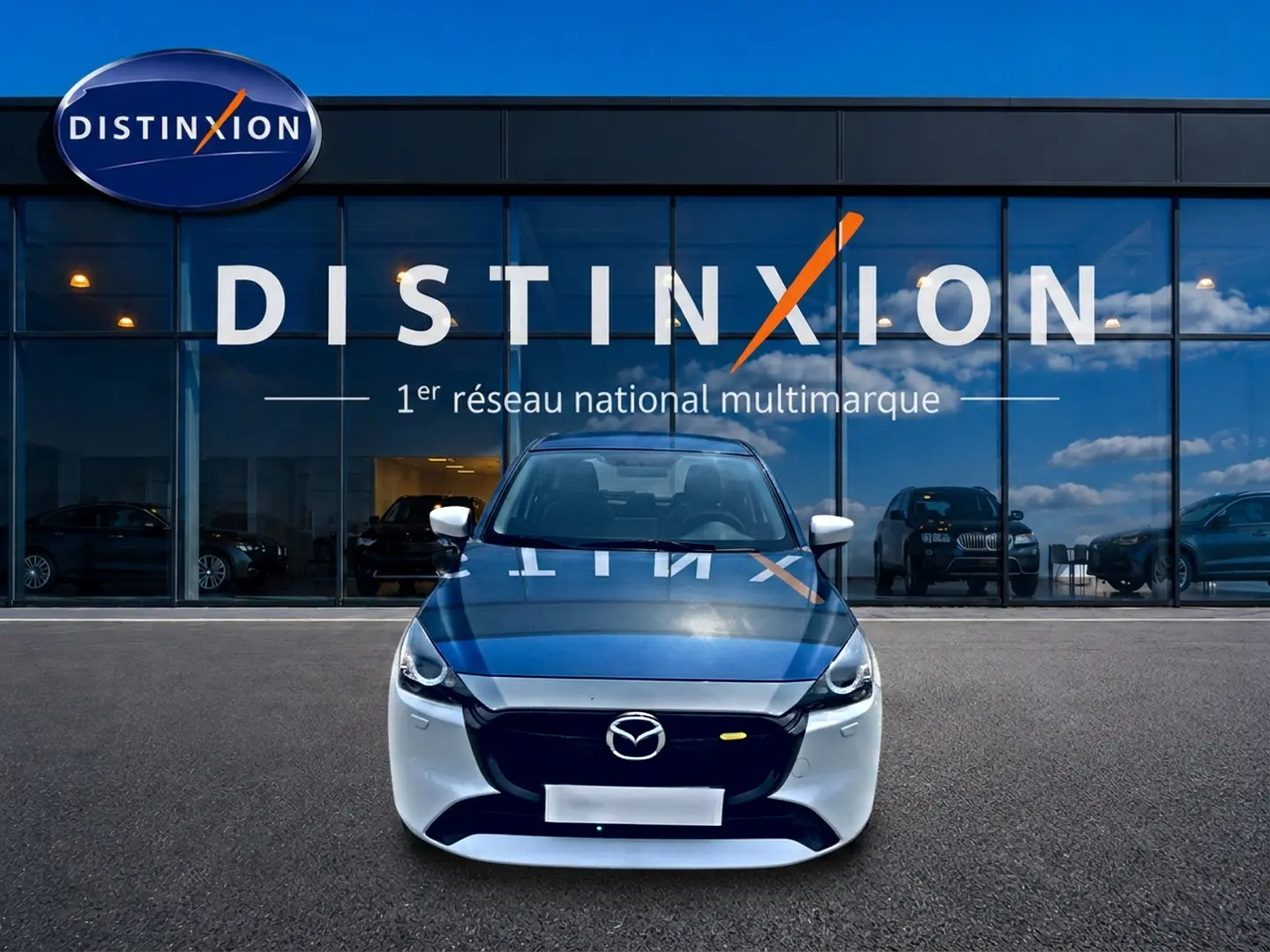 Vue avant d'une Mazda2 blanche 2024 avec calandre noire, stationnée devant une vitrine de concession multimarque.