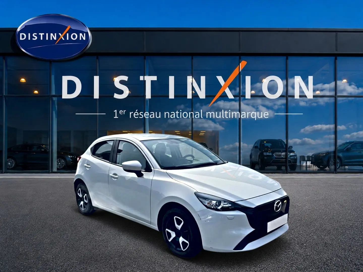 Vue 3/4 avant gauche d'une Mazda2 blanche 2024 avec calandre noire et jantes distinctives devant un showroom Distinxion.