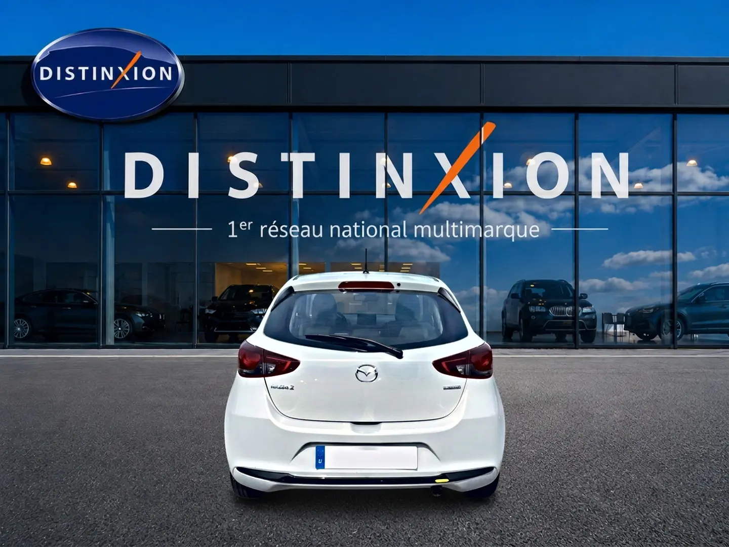 Vue arrière d'une Mazda2 blanche 2024 garée devant une vitrine avec logo Distinxion en arrière-plan.