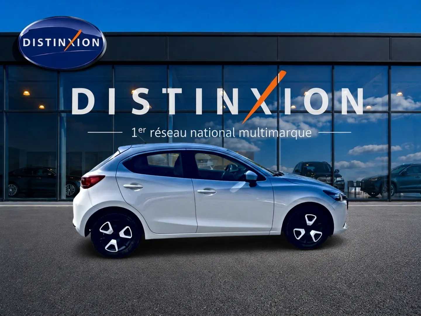 Vue de profil côté gauche d'une Mazda2 blanche 2024 garée devant une vitrine avec logo Distinxion.