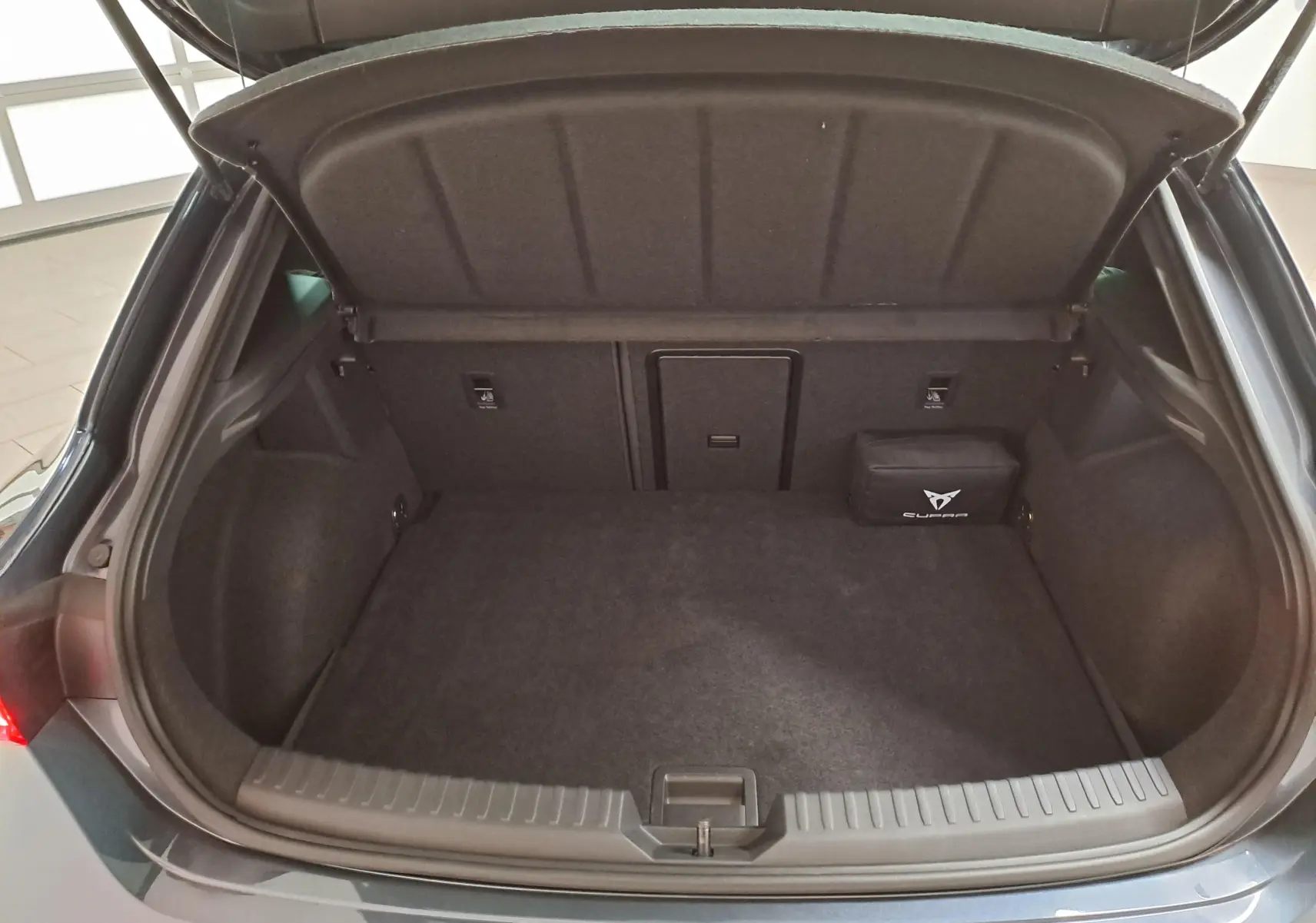 Coffre ouvert vu de l'arrière d'une CUPRA Leon gris magnétique 2023, avec tapis noir et trousse d'urgence.
