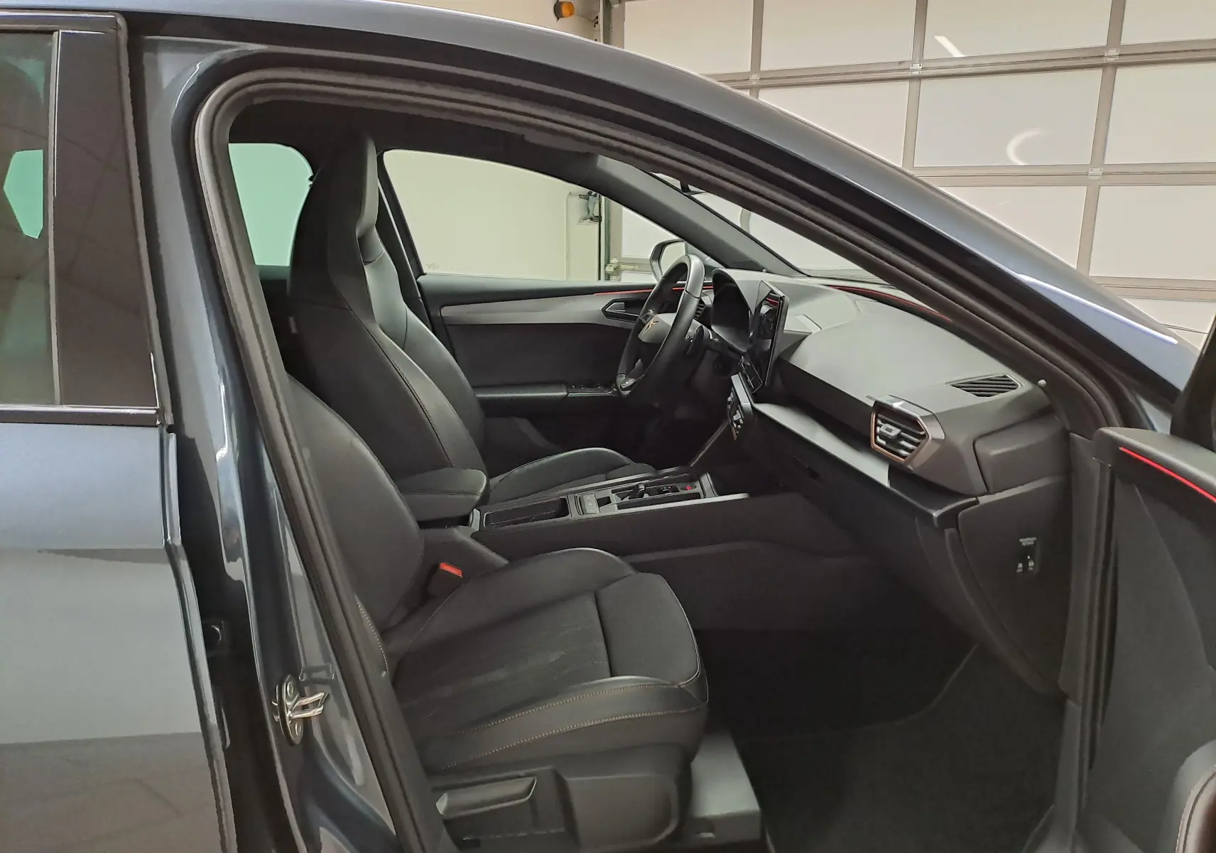 Vue intérieure côté gauche de la CUPRA Leon 1.5 TSI 150 DS7 V 2023 en gris magnétique, sièges noirs avec surpiqûres orange.