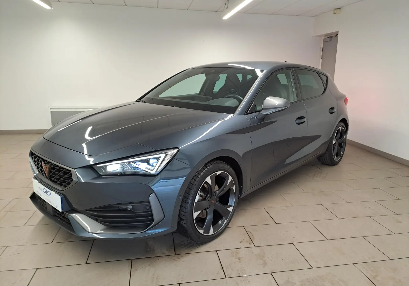 Vue 3/4 avant d'une CUPRA Leon 1.5 TSI 150 DS7 V gris magnétique avec jantes noires et phares allumés en intérieur.