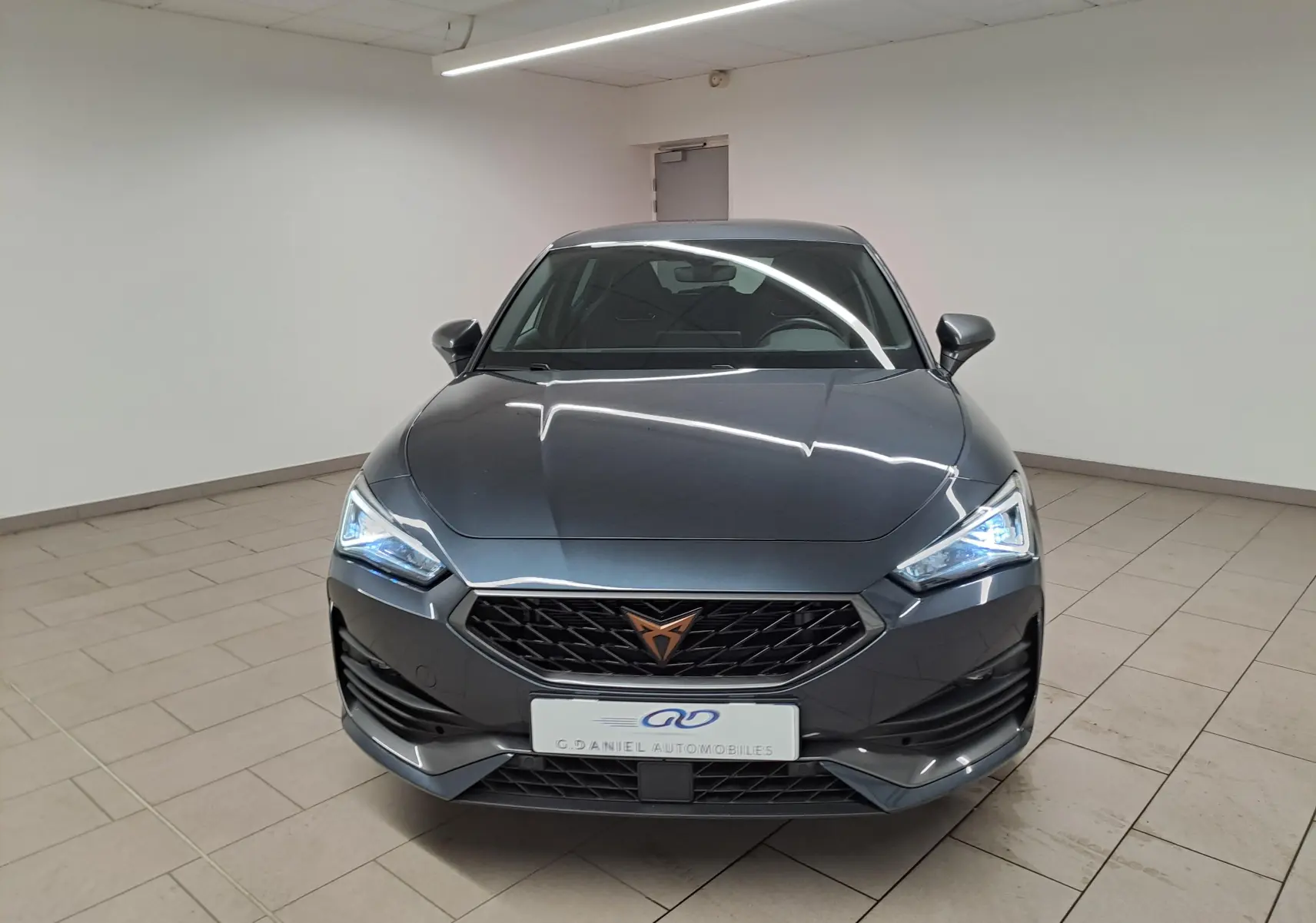 Vue frontale d'une CUPRA Leon 2023 gris magnétique avec calandre noire et logo cuivré, phares LED allumés en intérieur.