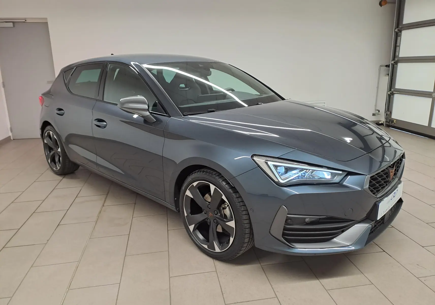 Vue 3/4 avant droit d'une CUPRA Leon 1.5 TSI 150 DS7 V gris magnétique avec jantes noires et détails sportifs.