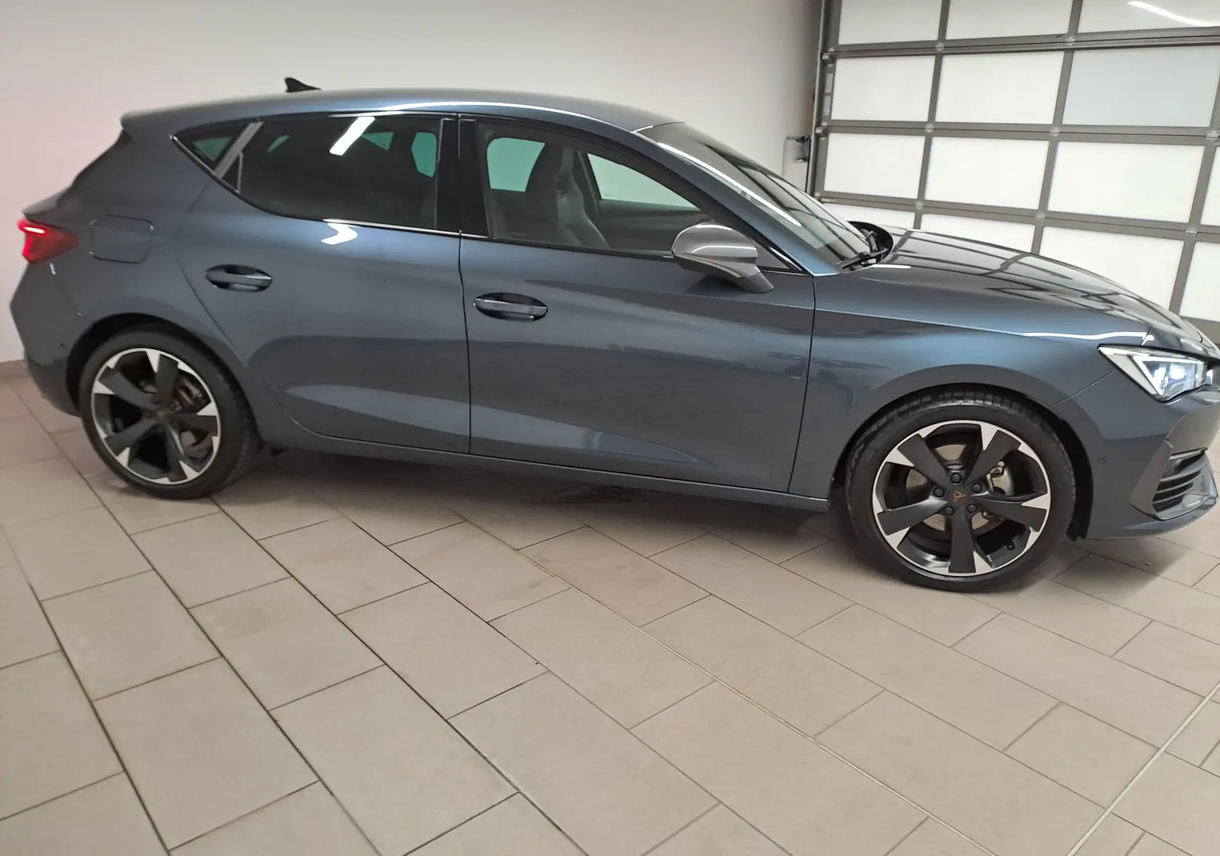 Vue de profil côté gauche d'une CUPRA Leon 2023 gris magnétique avec jantes noires et grises dans un garage.