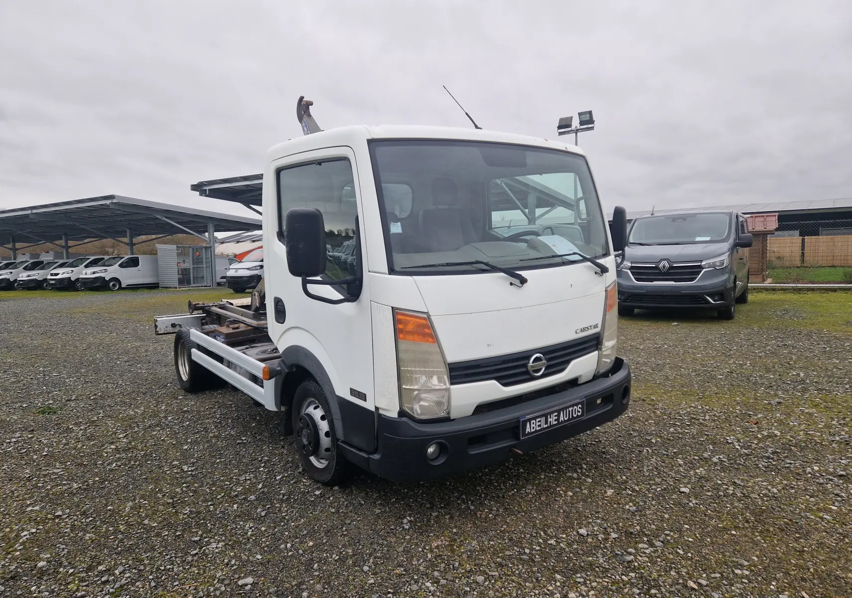 Vue 3/4 avant d'un Nissan Cabstar blanc 2010 avec châssis cabine et bras polybenne visible sur un terrain gravillonné.