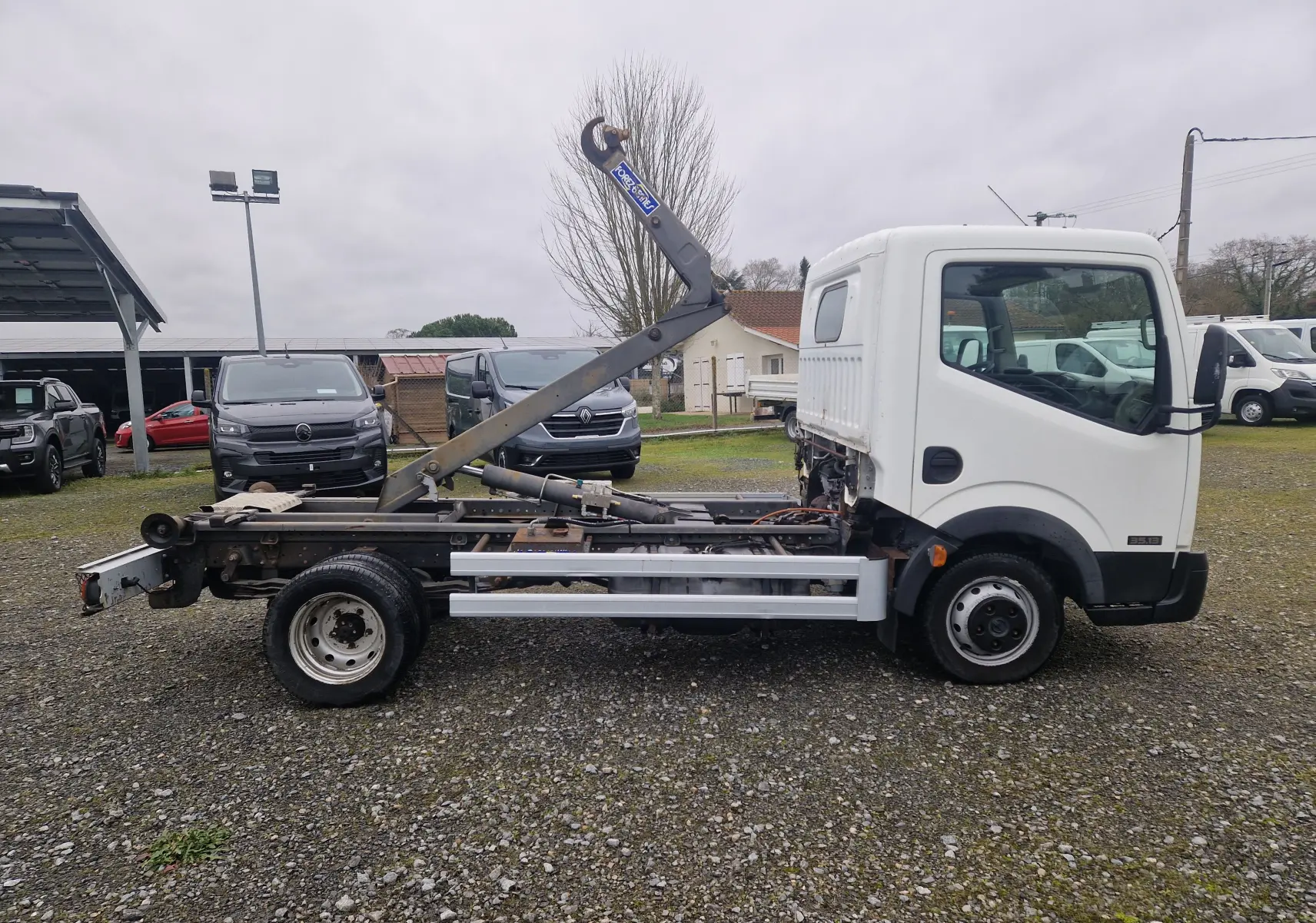 Vue de profil côté droit d’un Nissan Cabstar blanc 2010 avec châssis cabine et bras polybenne relevé sur un terrain extérieur.