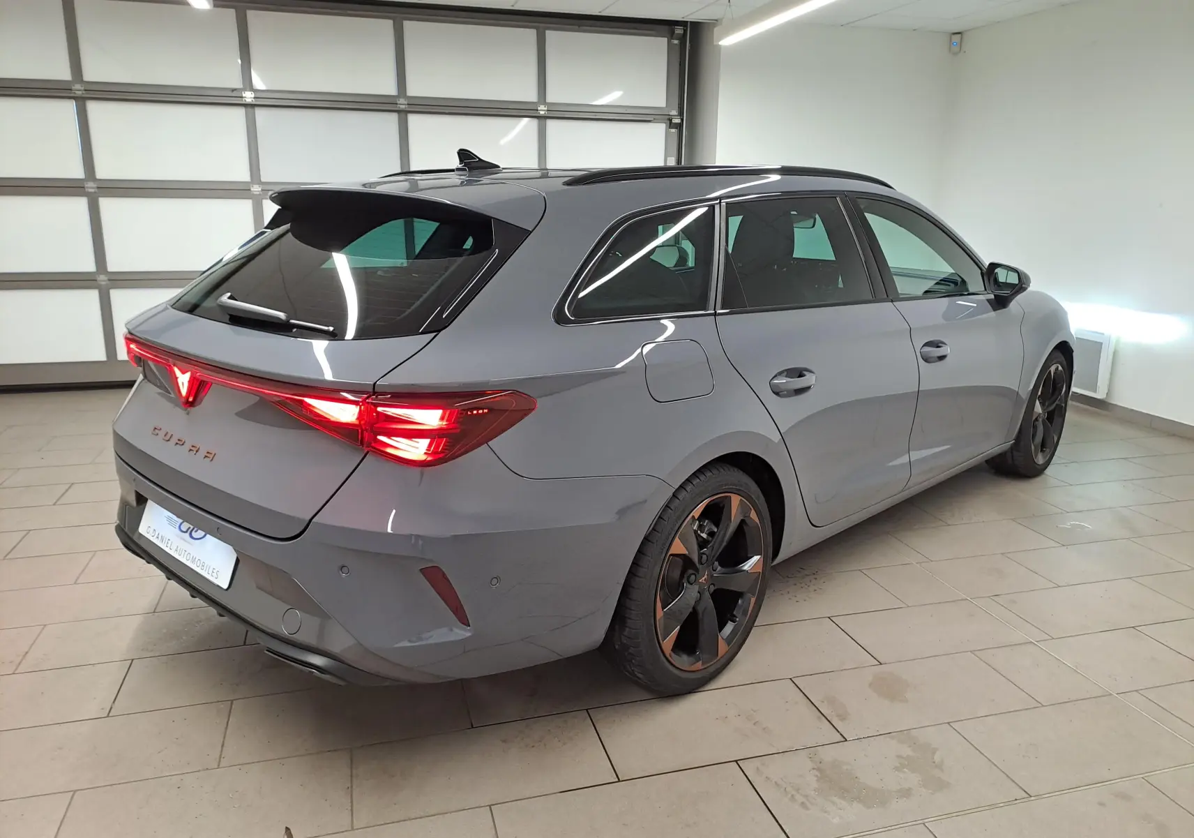 Vue 3/4 arrière droite d'une CUPRA Leon ST gris Graphène avec jantes cuivrées et toit ouvrant panoramique en intérieur showroom.