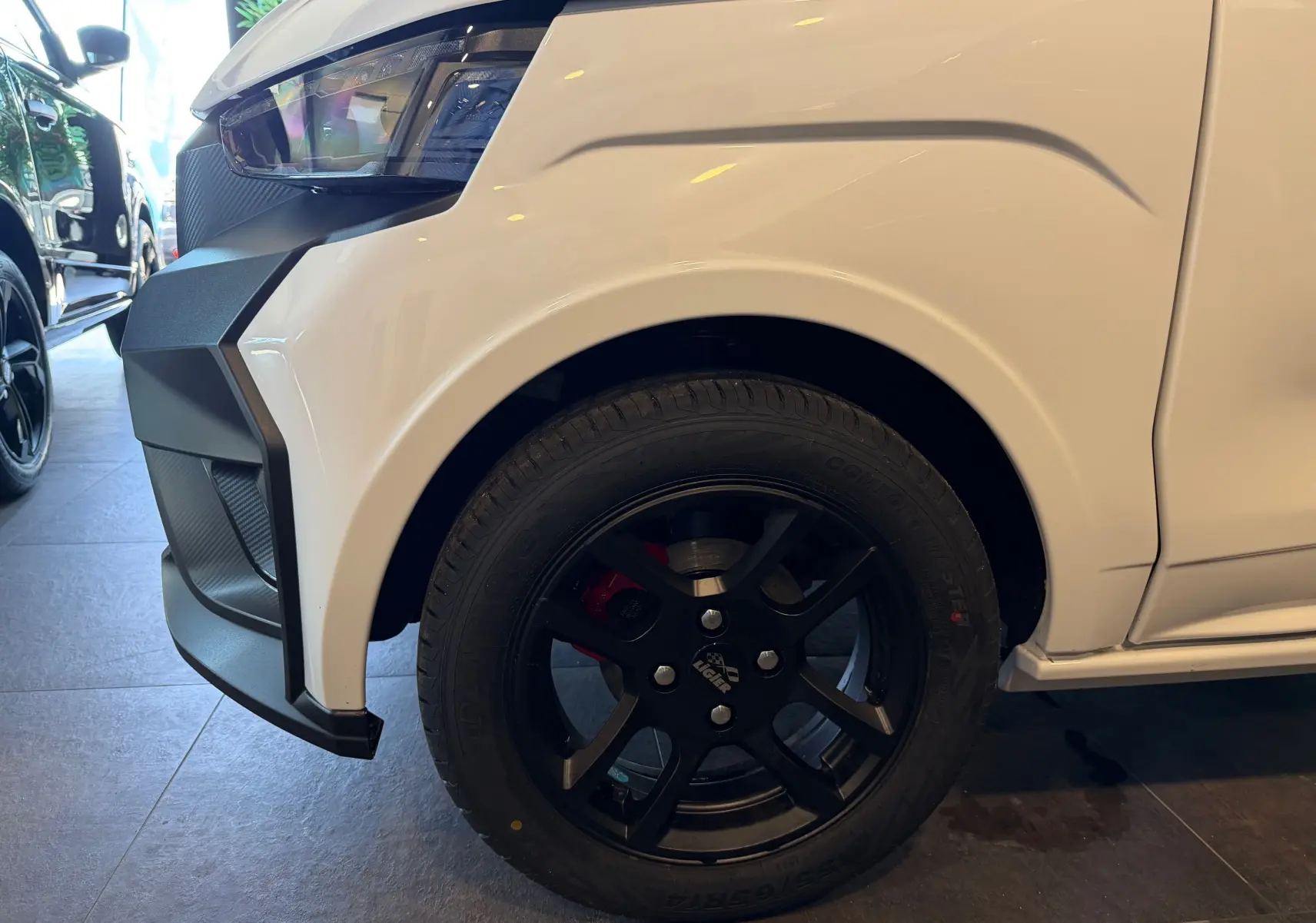 Gros plan sur la roue avant droite noire et l’aile blanche glacier d’une Ligier JS50 Select Revo D+ 2026 en intérieur showroom.