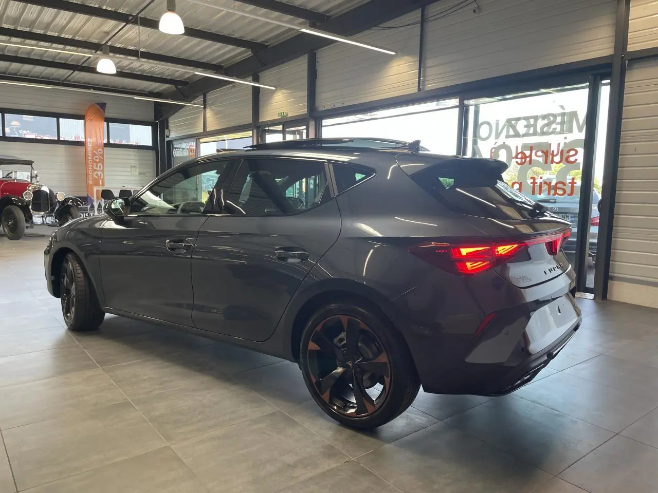 Vue 3/4 arrière droite d'une CUPRA Leon gris magnétique avec jantes cuivrées et feux arrière allumés en intérieur showroom.