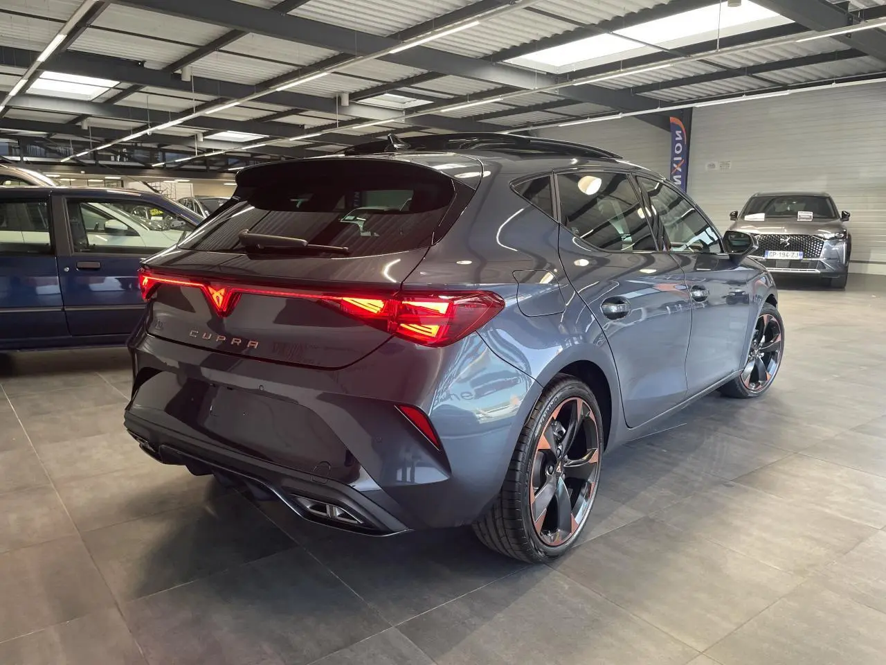 Vue 3/4 arrière droite d'une CUPRA Leon gris magnétique avec jantes cuivrées et feux arrière allumés en showroom.